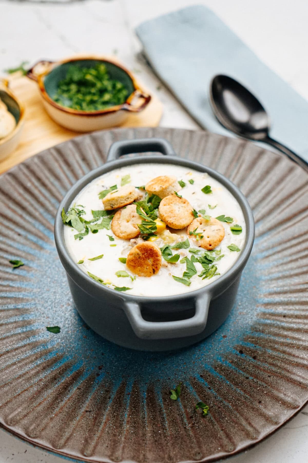 A bowl of creamy soup garnished with crackers and chopped herbs, served on a plate with a napkin and a small bowl of additional herbs in the background.