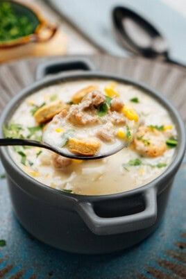 A bowl of creamy sausage corn soup garnished with parsley, corn, and oyster crackers, with a spoon lifting a portion above the bowl.