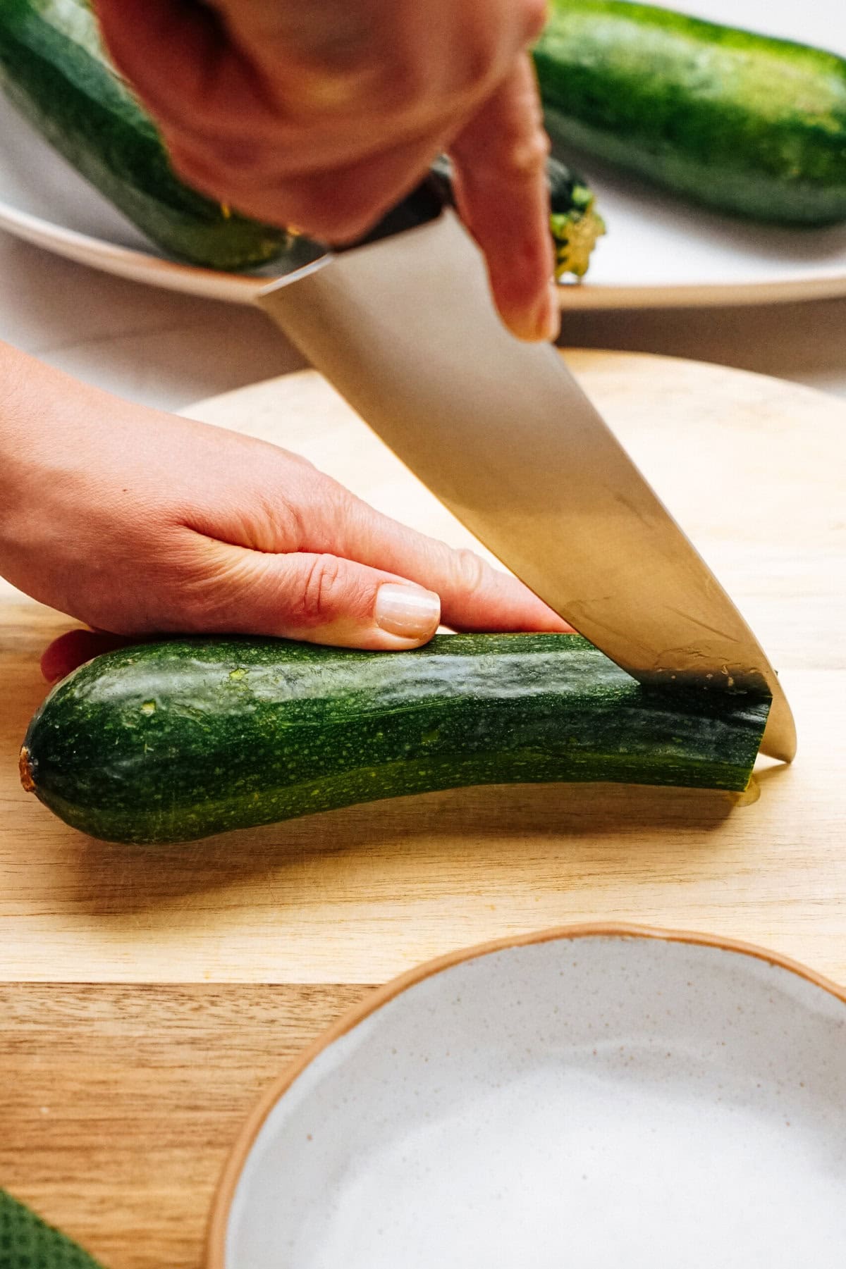 A close-up of a hand slicing a green zucchini on a wooden cutting board with a large knife. A white plate and partially visible zucchini are in the background.