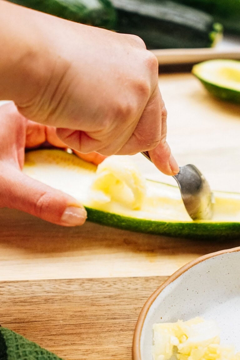 A person uses a spoon to scoop out the seeds from a halved zucchini on a wooden cutting board.