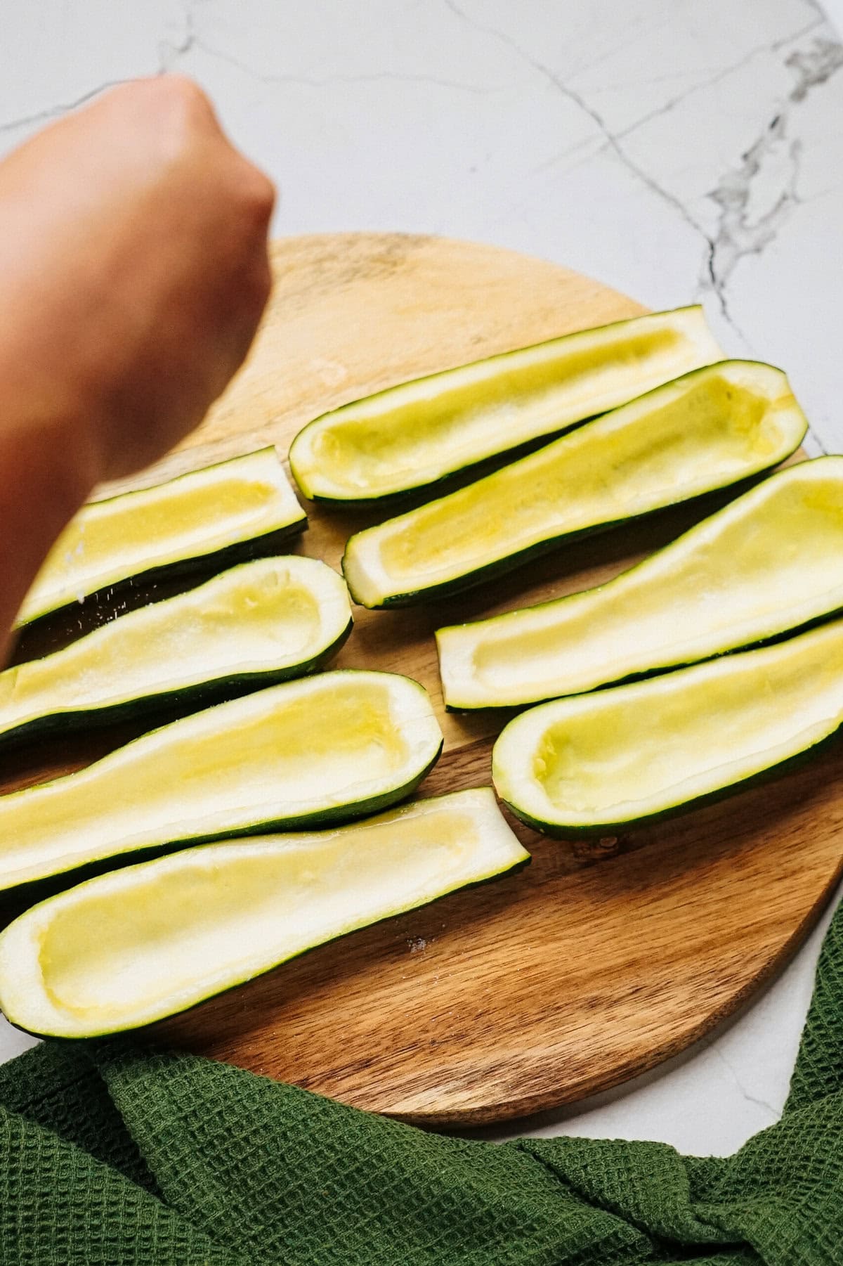 A hand sprinkles seasoning onto eight hollowed-out zucchini halves arranged on a round wooden cutting board, with a green cloth partially visible at the bottom.