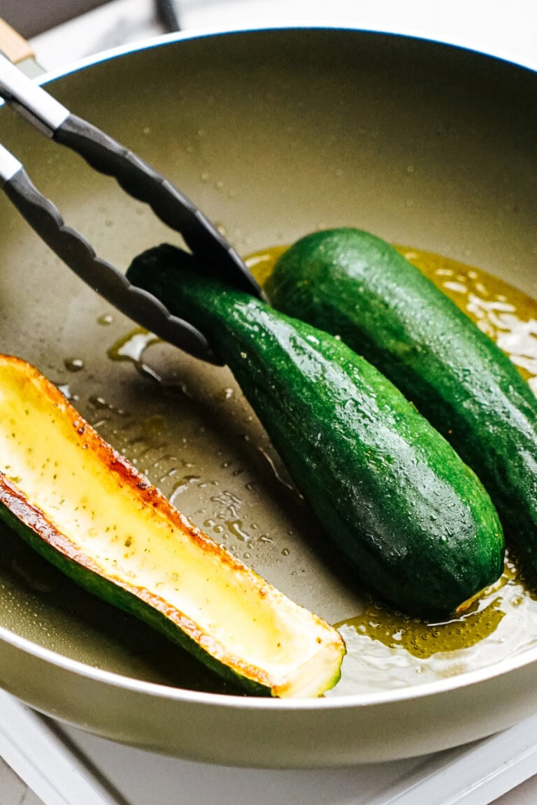 Cooking zucchinis in a frying pan, with one zucchini half being turned over using tongs in hot oil.