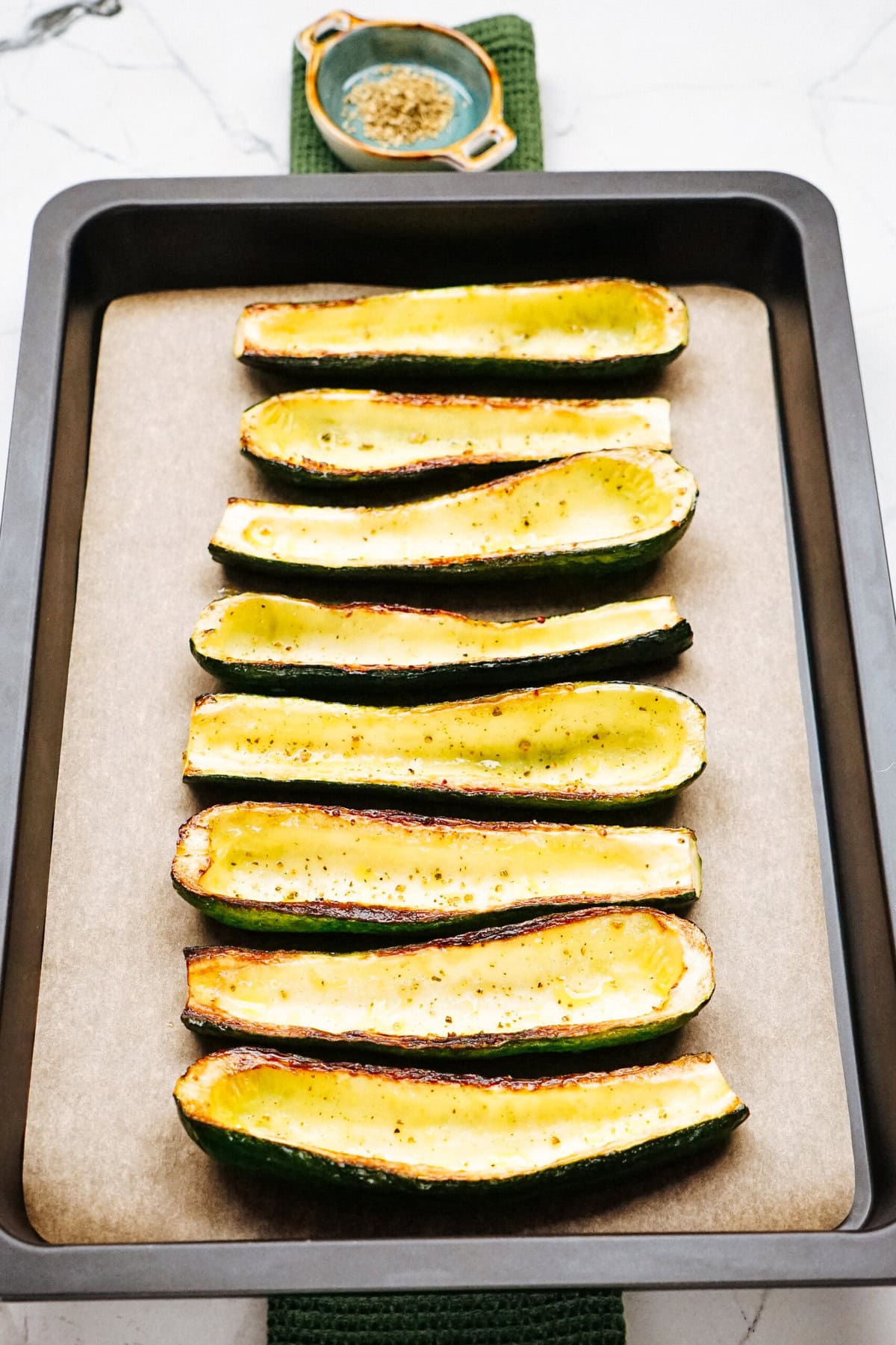 Baking tray with eight zucchini boats placed on parchment paper, seasoned lightly. A small dish with seasoning is in the background.