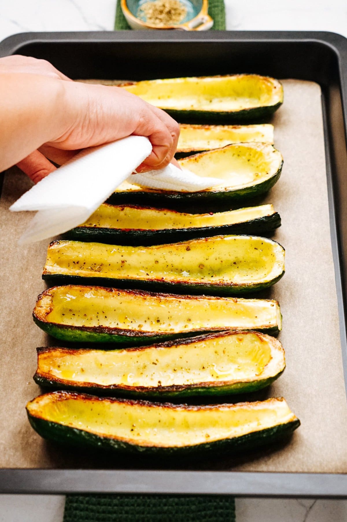 A hand uses a paper towel to blot moisture from halved and seasoned zucchinis arranged in a baking tray lined with parchment paper.