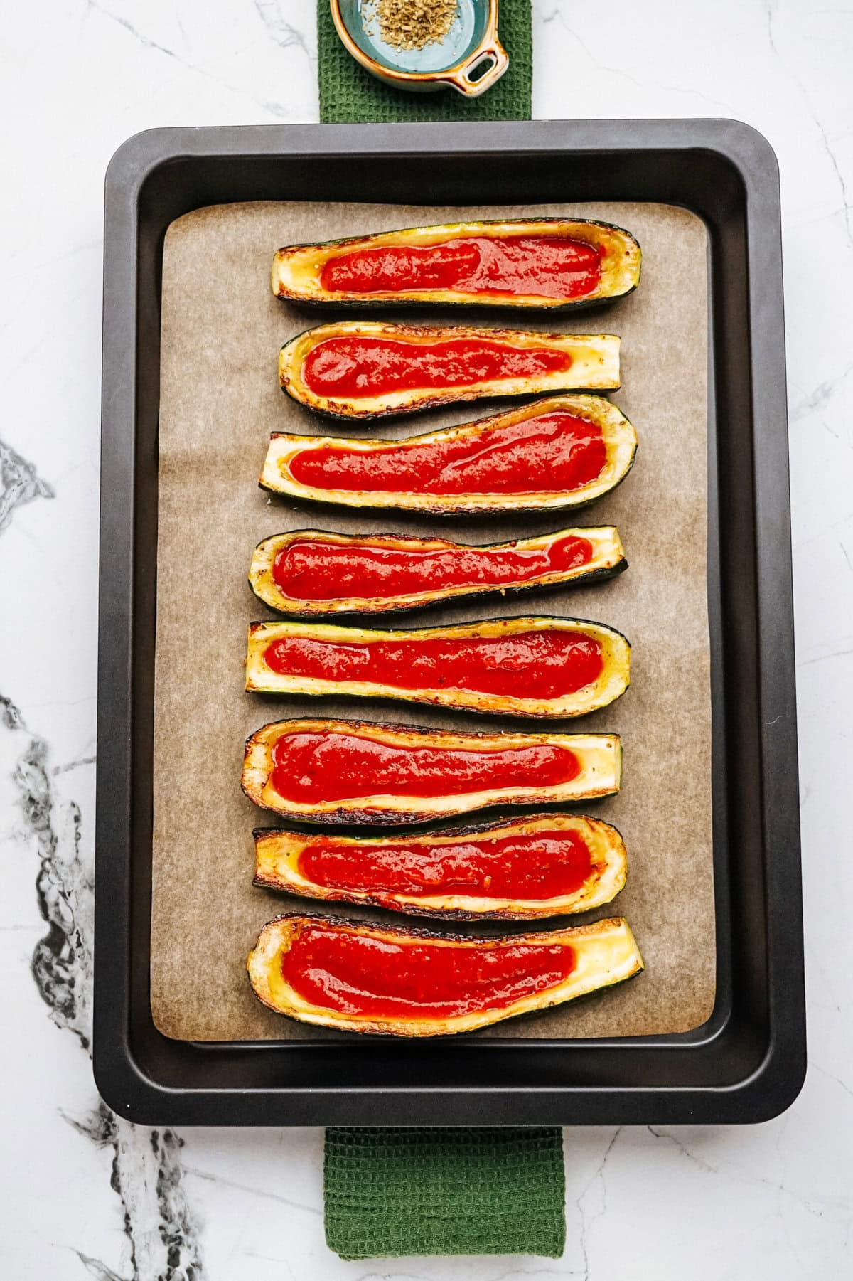 A baking tray with eight halves of zucchini filled with marinara sauce placed neatly in two rows on parchment paper.