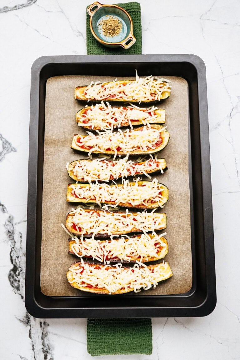 A baking tray with eight zucchini halves topped with tomato sauce and shredded cheese, placed on parchment paper. A small bowl with seasoning is above the tray on a green cloth.