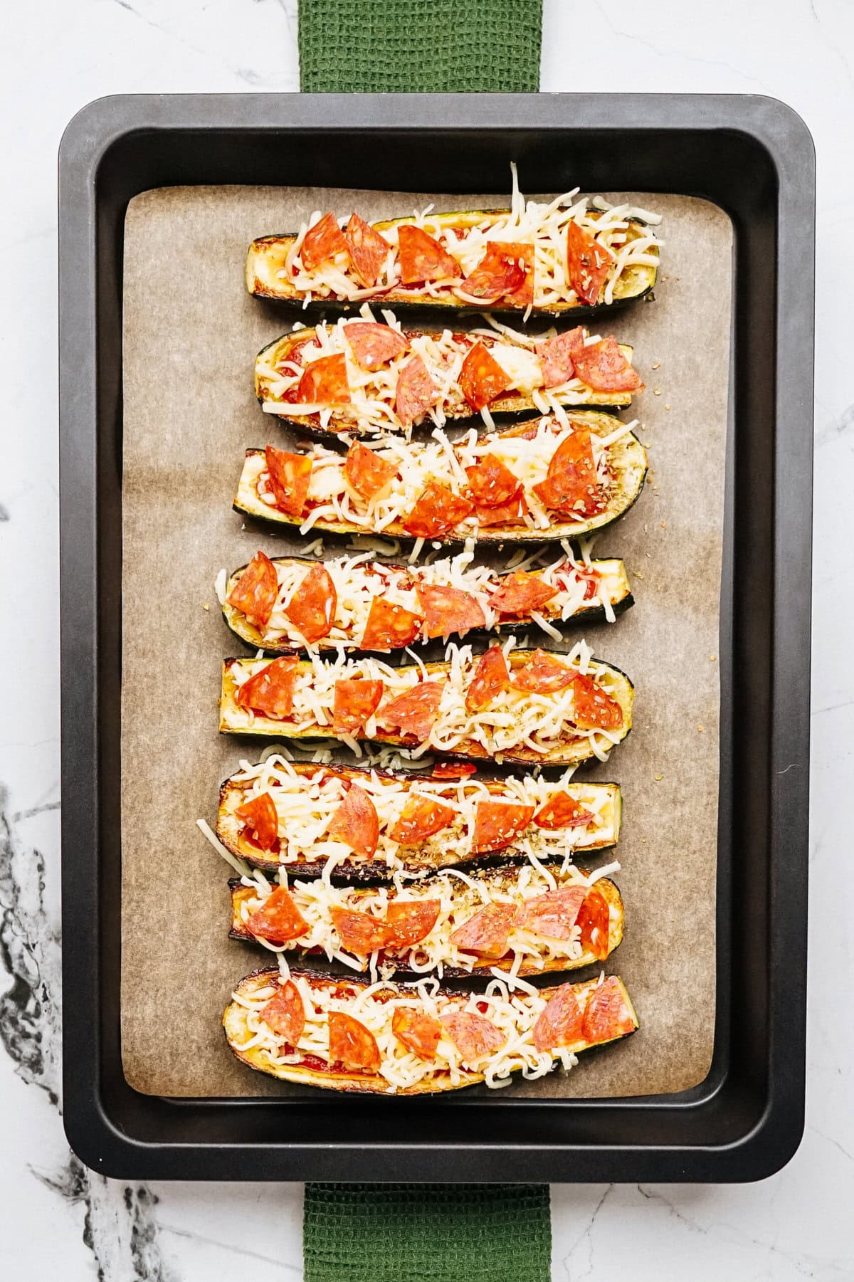 A baking tray with six halved zucchinis topped with shredded cheese and pepperoni, ready to be baked.