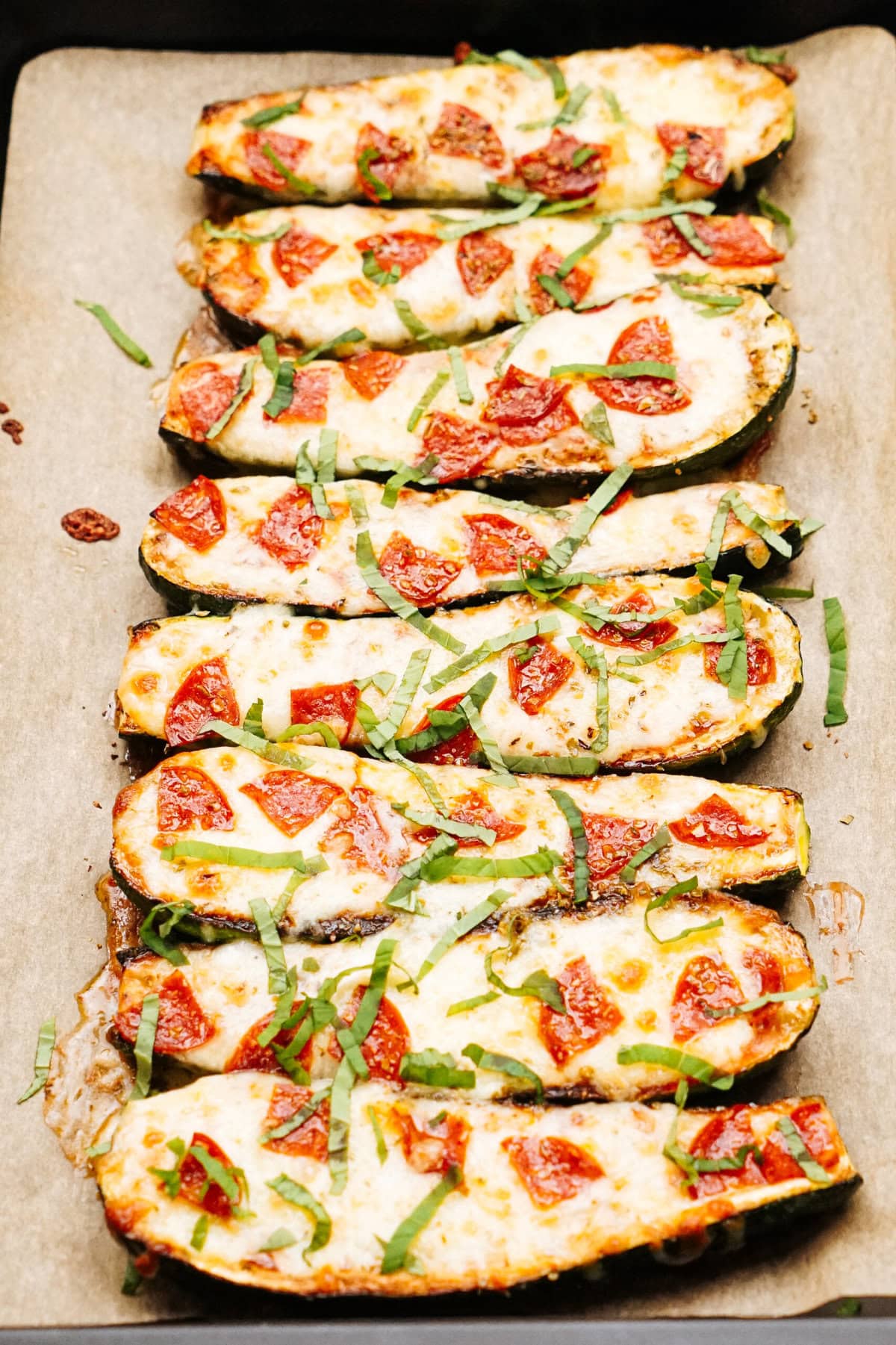 Four zucchini boats topped with melted cheese, tomato pieces, and fresh herbs on a parchment