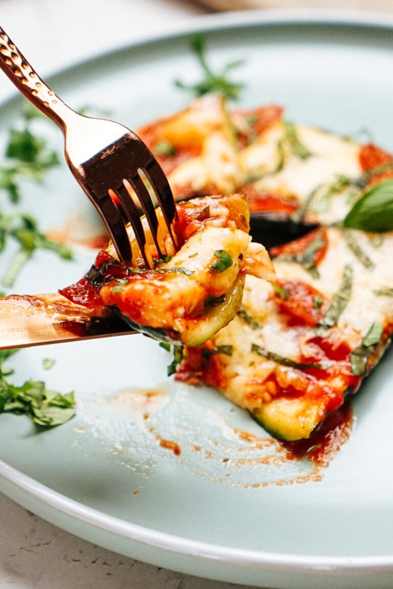 A fork holding a bite of vegetable lasagna above a plate with additional lasagna pieces garnished with fresh herbs.