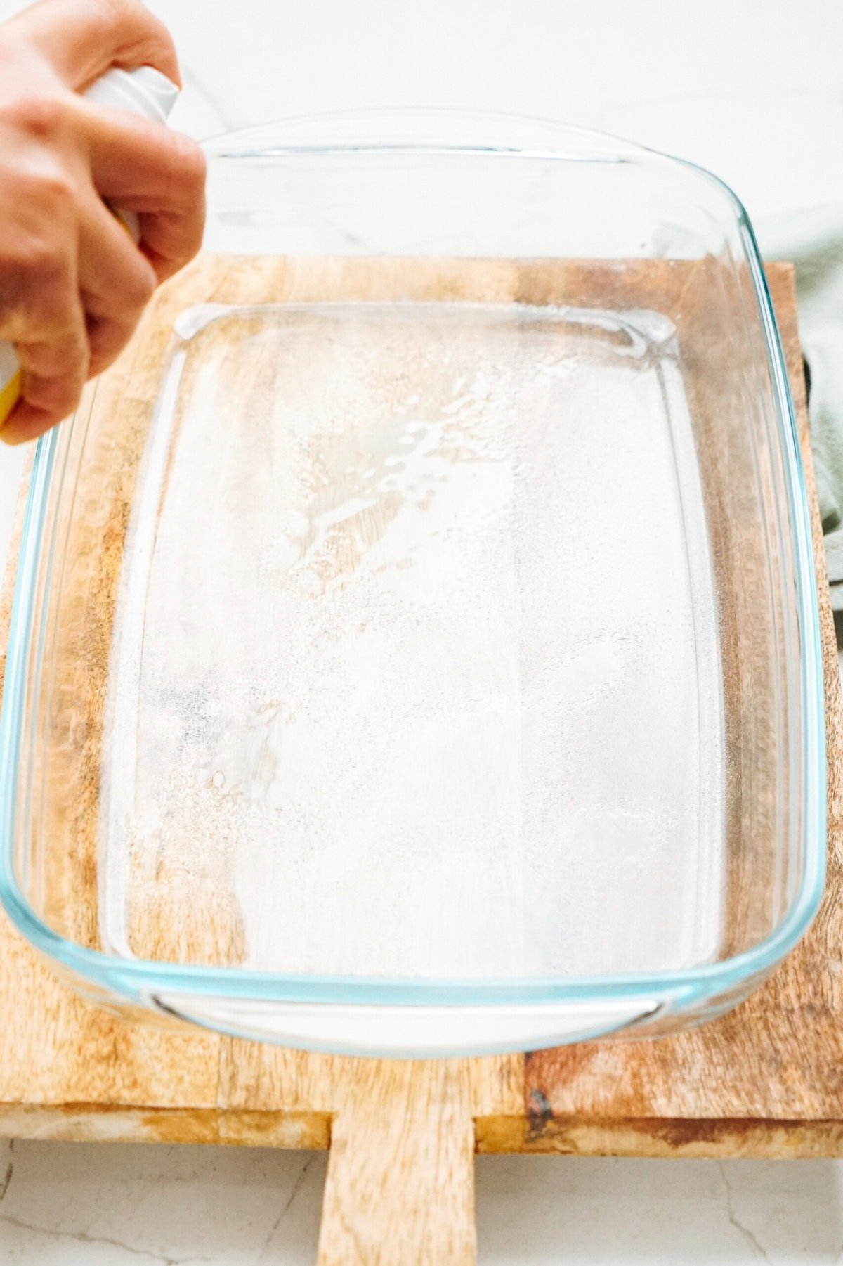 A hand sprays a clear glass baking dish with oil, preparing it for the upcoming ingredients of a delicious Zucchini Pizza Casserole, all set on a wooden surface.