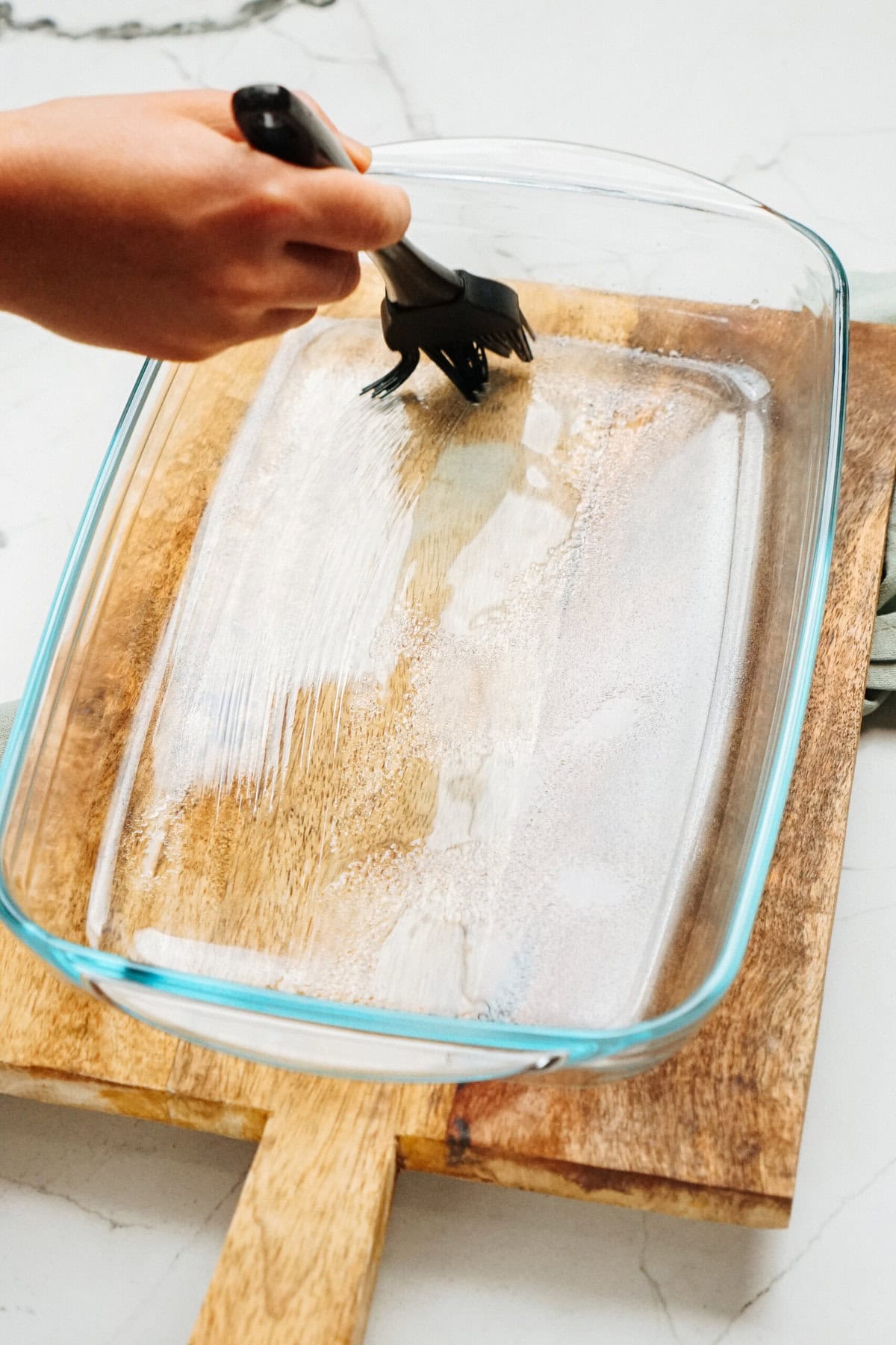 A hand uses a brush to apply oil or butter to a clear rectangular baking dish placed on a wooden cutting board, prepping it for the delicious Zucchini Pizza Casserole.