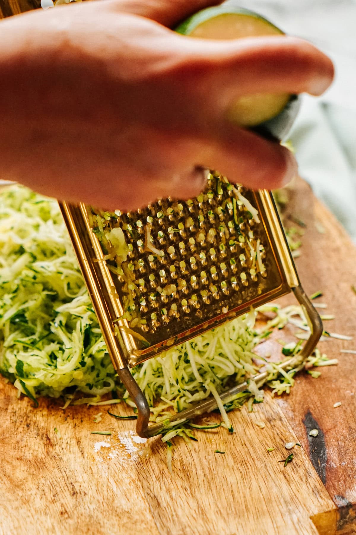 A person grating zucchini on a metal grater over a wooden surface, preparing ingredients for a delicious Zucchini Pizza Casserole, with a pile of grated zucchini below.