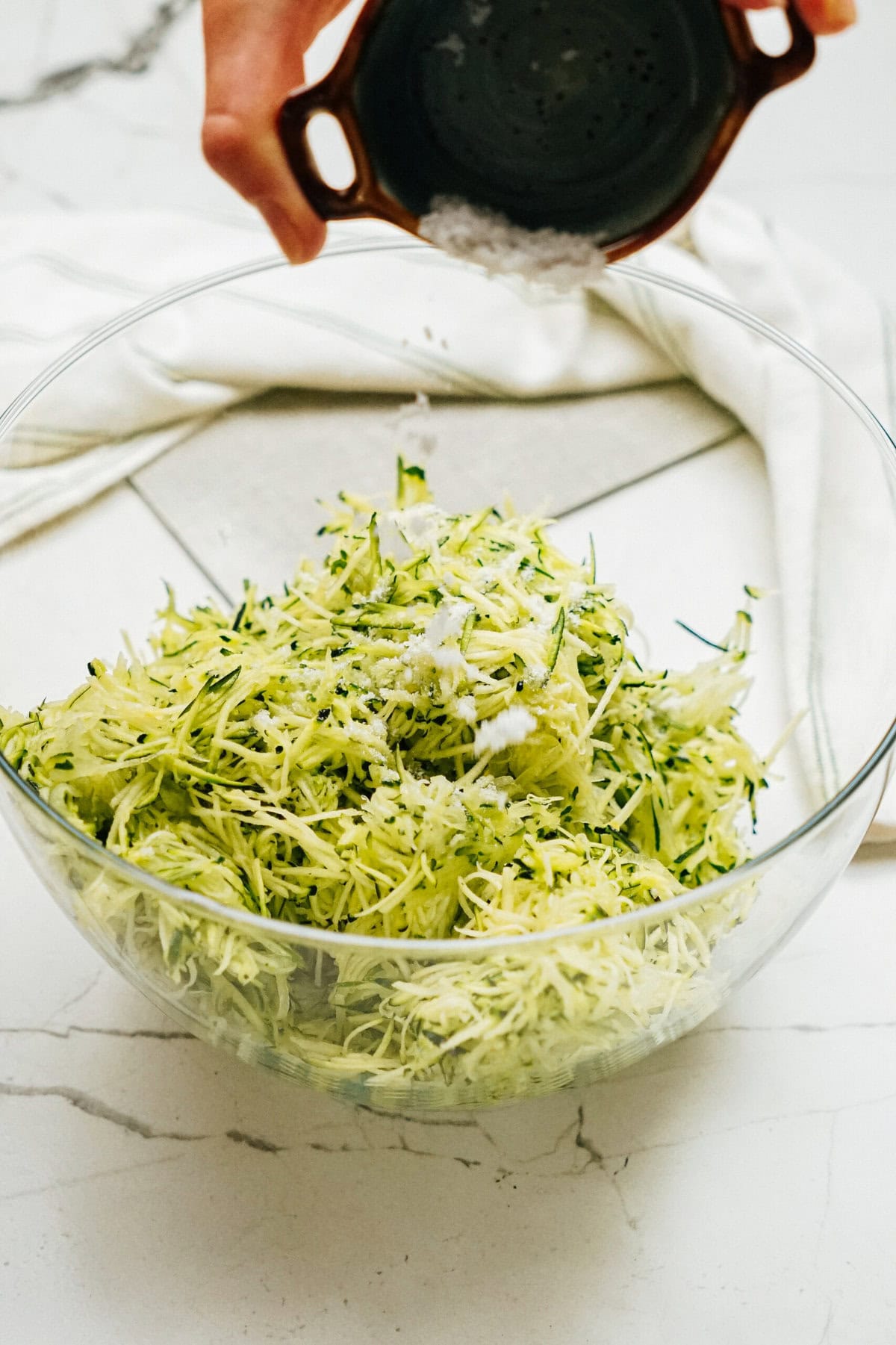 A hand is sprinkling salt from a small, dark bowl onto a large, clear glass bowl filled with shredded zucchini on a white countertop, preparing for a delicious Zucchini Pizza Casserole.