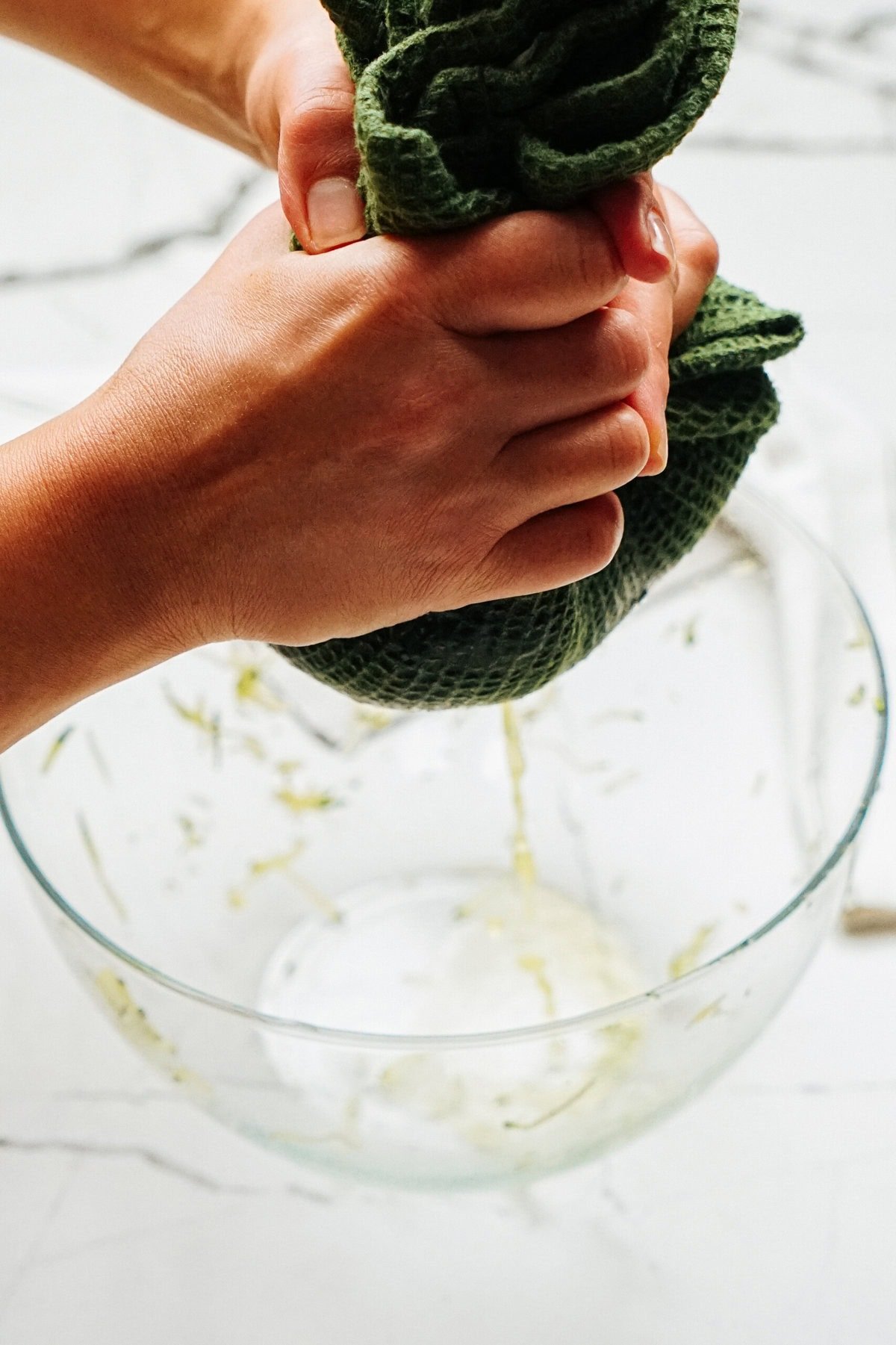 Hands squeezing liquid from a green cloth over a glass bowl on a marble surface, prepping ingredients for the zucchini pizza casserole.