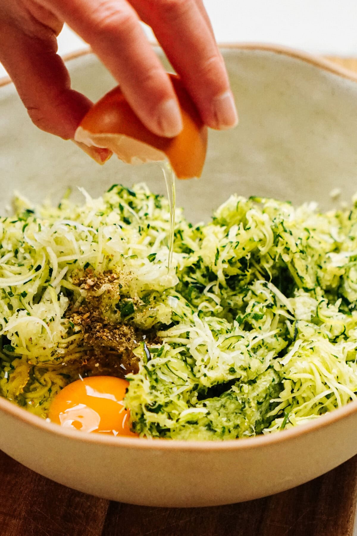 A hand cracks an egg over a bowl containing shredded zucchini and spices, preparing the base for a delicious Zucchini Pizza Casserole.