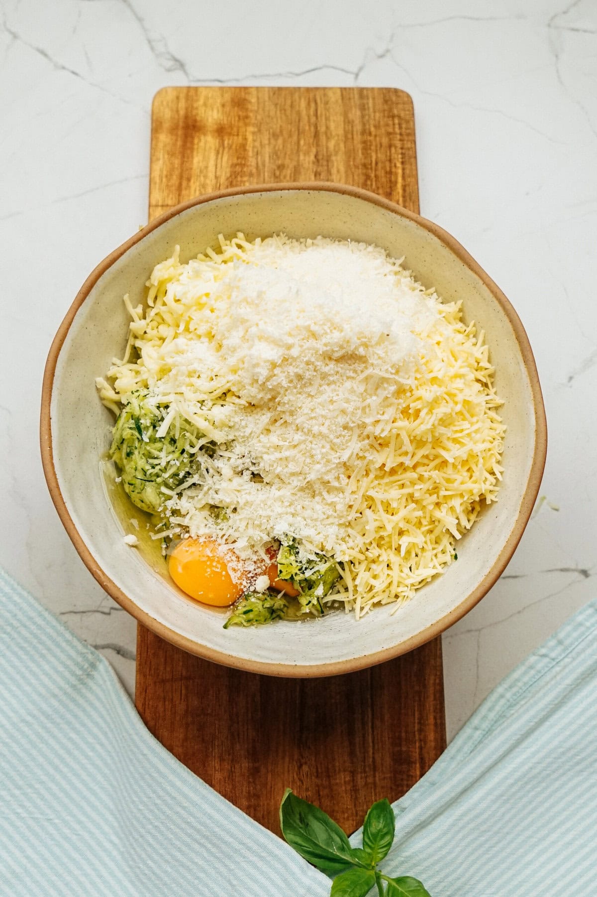 A bowl on a wooden board filled with grated cheese, eggs, and herbs sits ready for the Zucchini Pizza Casserole, accompanied by a faint blue-striped cloth and a basil sprig nearby.