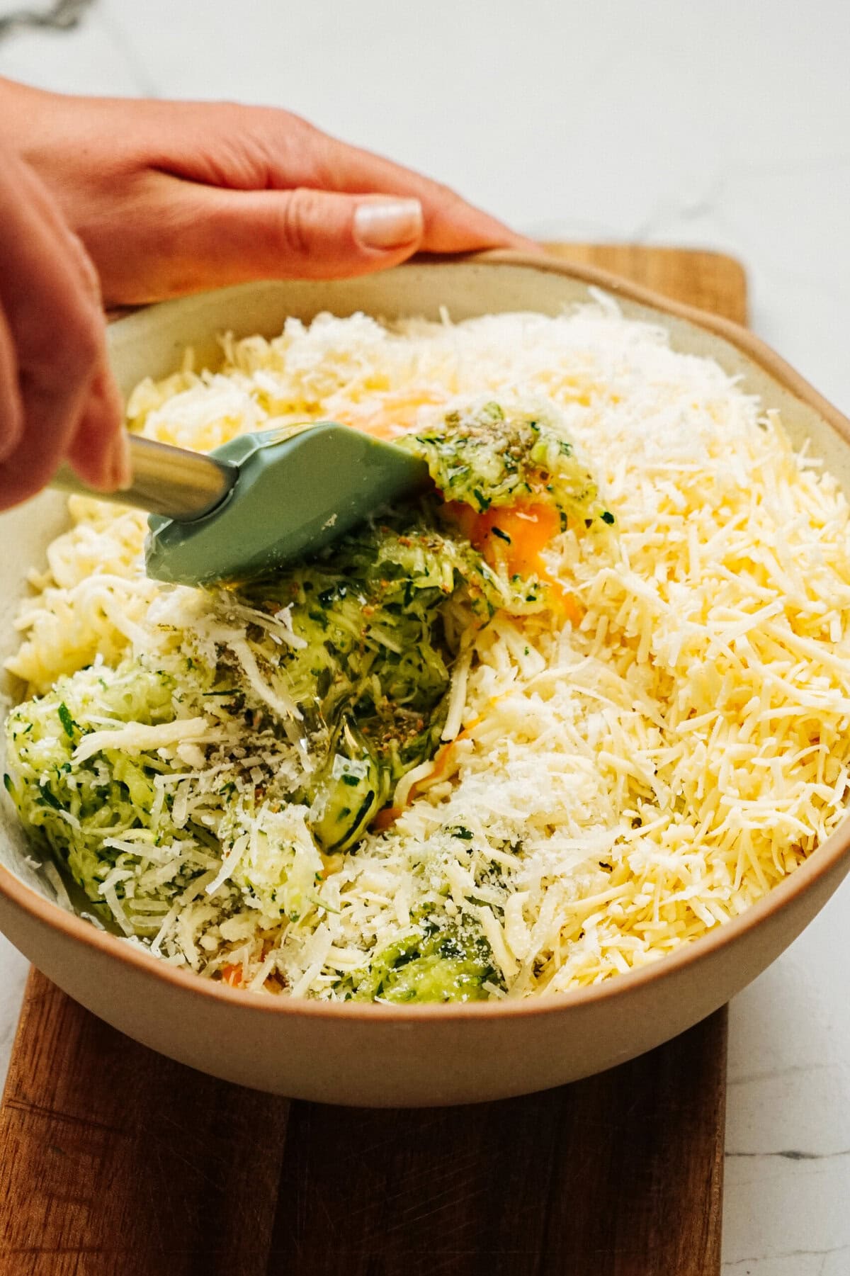 A person mixing grated zucchini, cheese, and other ingredients in a bowl with a spatula on a wooden surface, preparing the base for their delicious Zucchini Pizza Casserole.