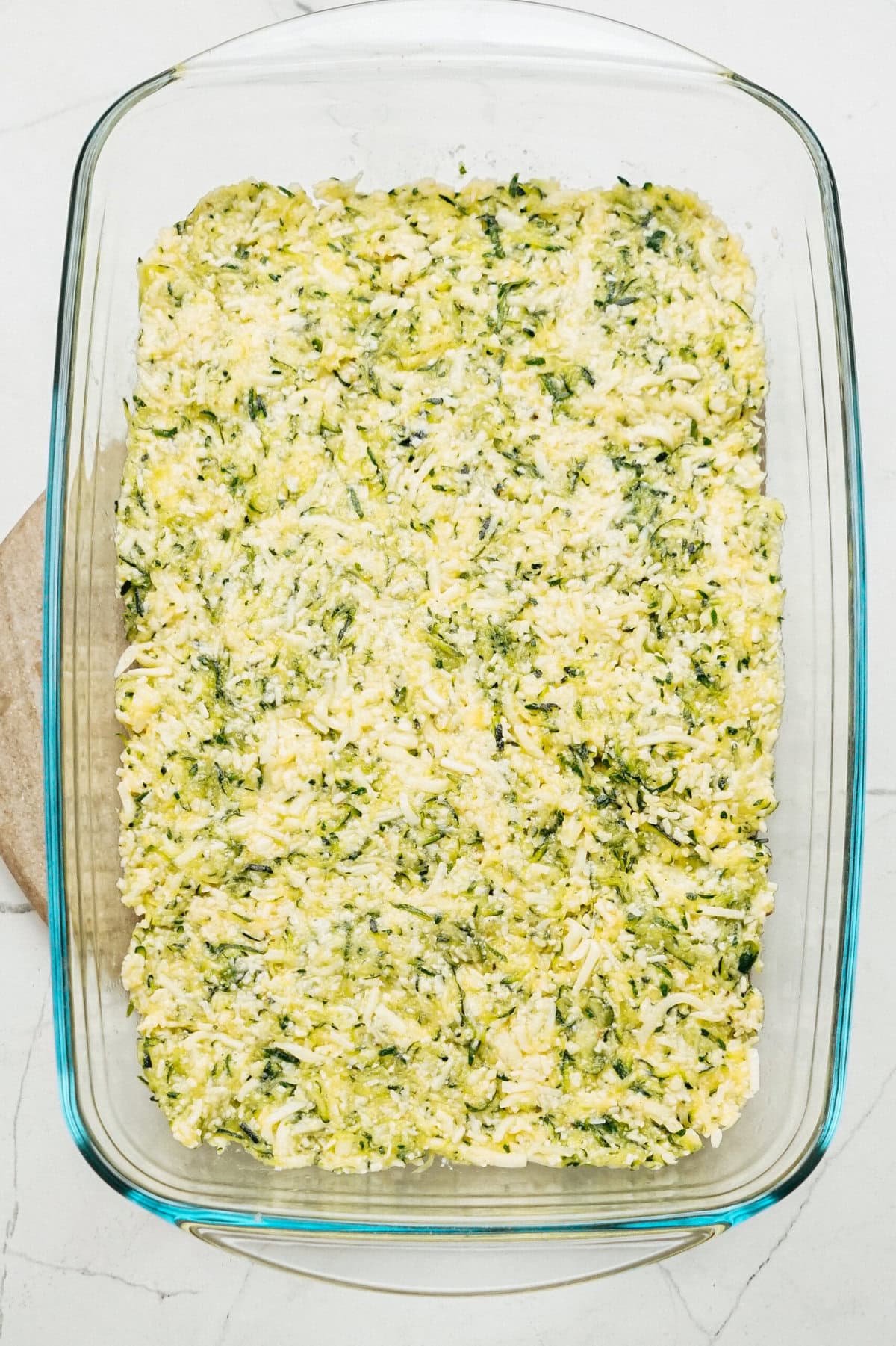 A clear glass baking dish filled with a mixture of shredded zucchini and cheese, cleverly prepared for a delicious Zucchini Pizza Casserole. Ready to be cooked to perfection.