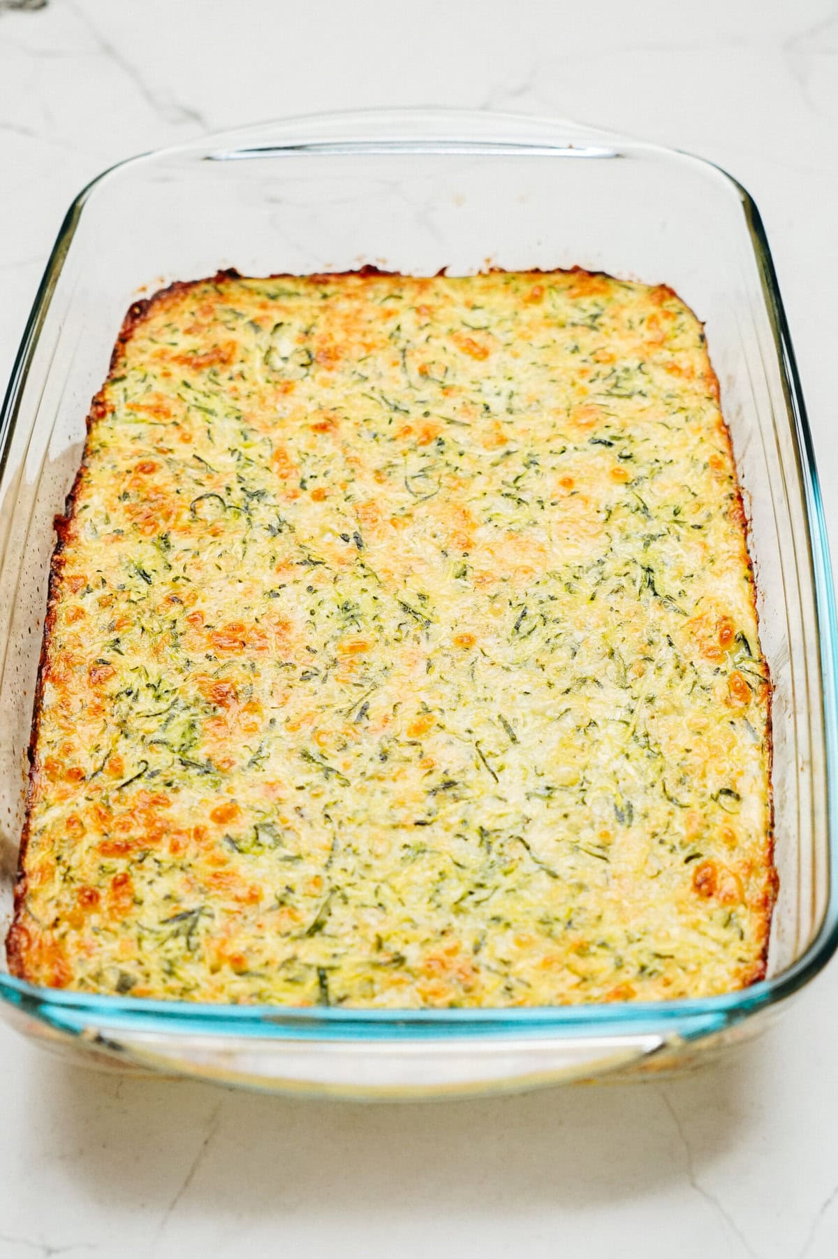 A baked casserole in a rectangular glass dish, featuring a golden-brown surface with green and white flecks of mixed vegetables and cheese, reminiscent of a delightful zucchini pizza casserole.