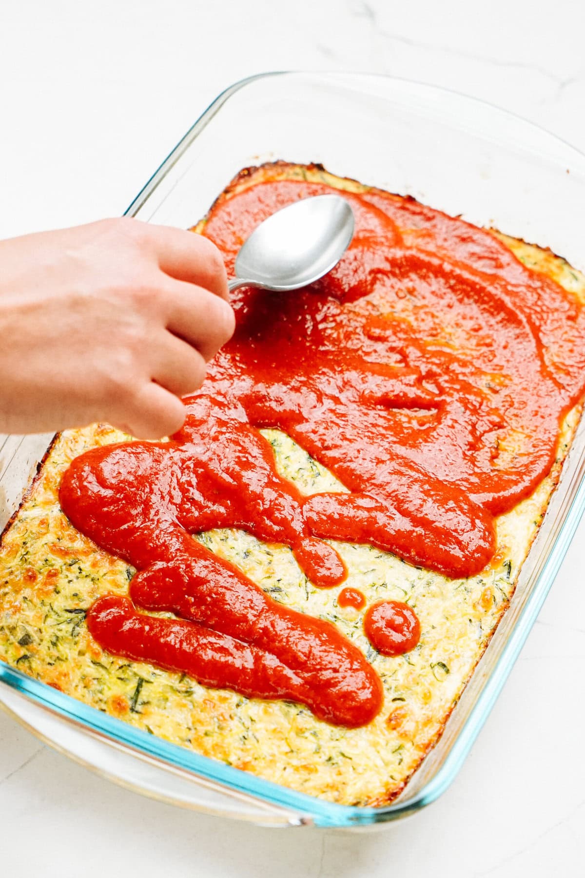 A hand spreads tomato sauce over a rectangular Zucchini Pizza Casserole in a glass dish using a spoon.