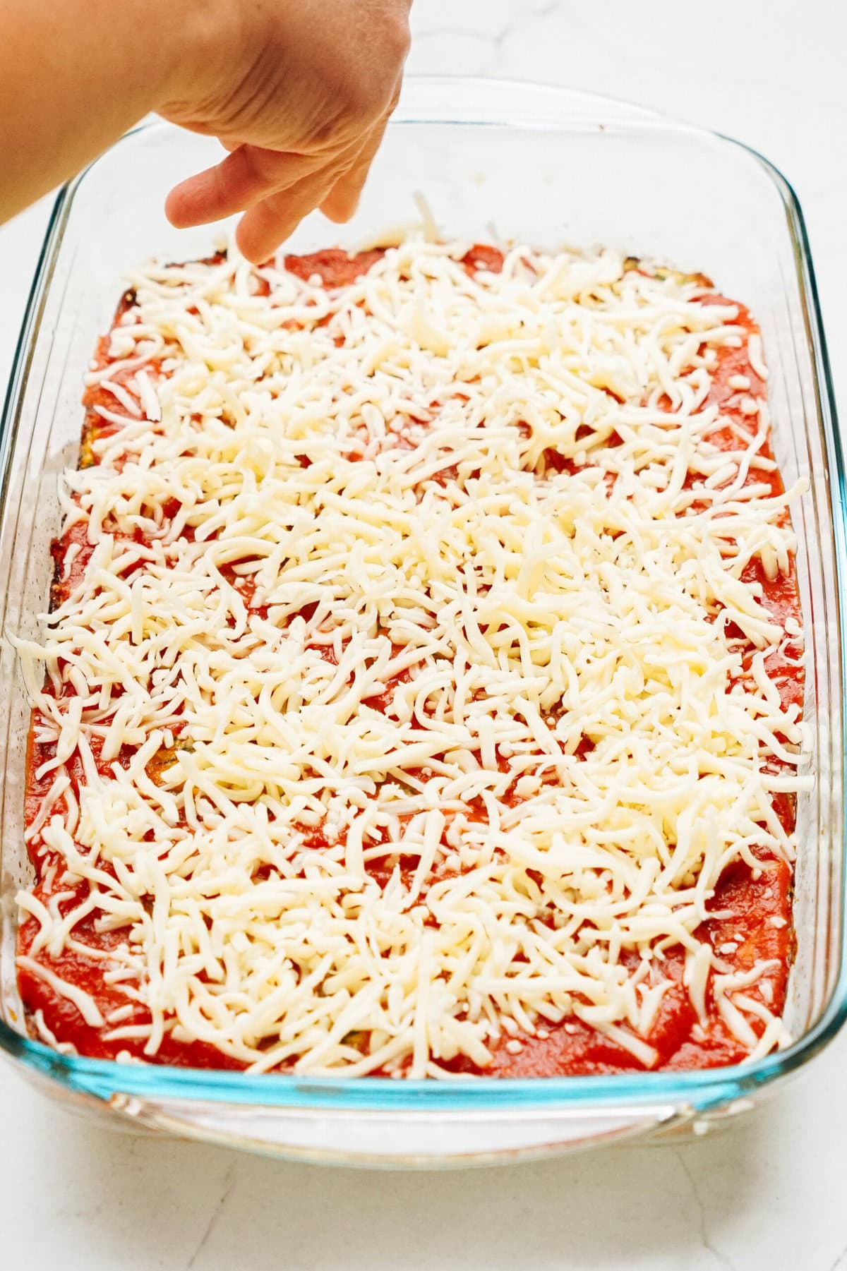 A hand sprinkling cheese over a dish of lasagna in a glass baking dish, turning it into a mouthwatering zucchini pizza casserole.