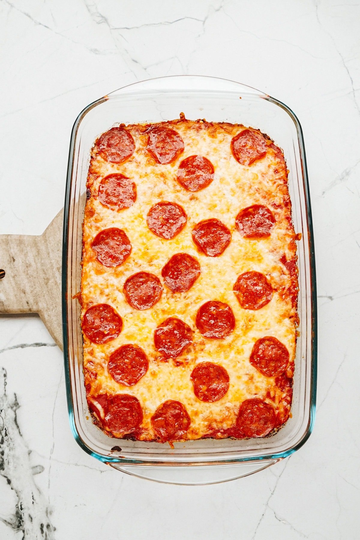 A glass baking dish filled with a delicious Zucchini Pizza Casserole, topped with pepperoni and cheese, sits on a marble countertop.