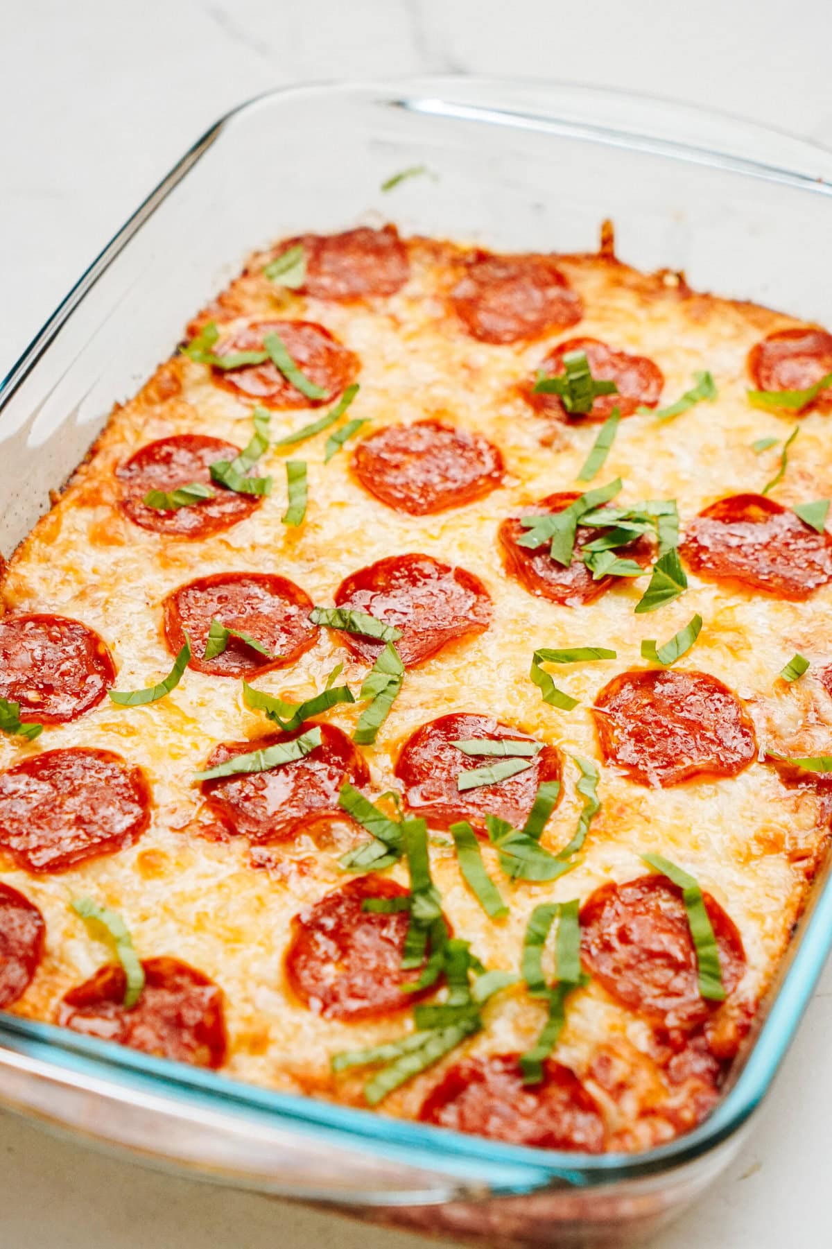 A baked Zucchini Pizza Casserole with melted cheese, sliced pepperoni, and chopped basil in a glass dish.