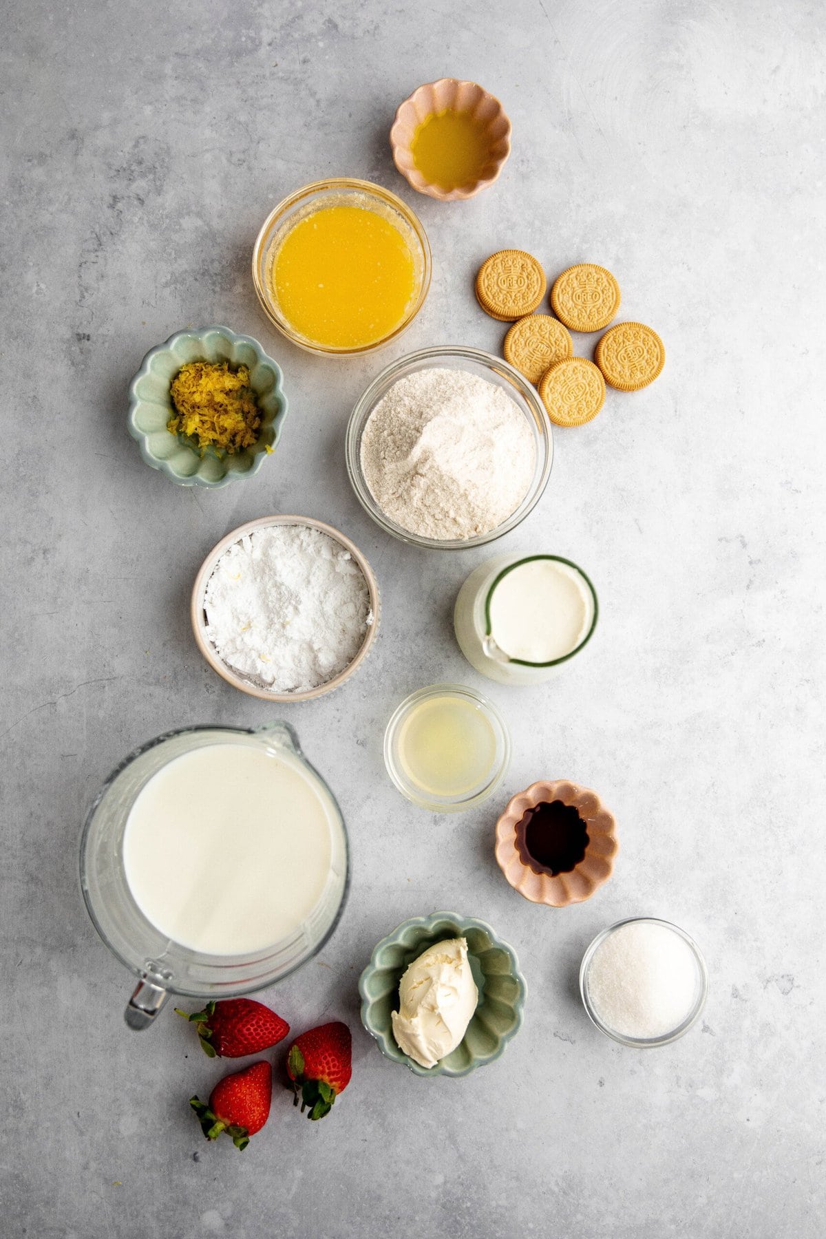 A flat lay of baking ingredients including flour, sugar, butter, vanilla extract, lemon zest, powdered sugar, cookies, milk, strawberries, and cream cheese, arranged on a gray surface.