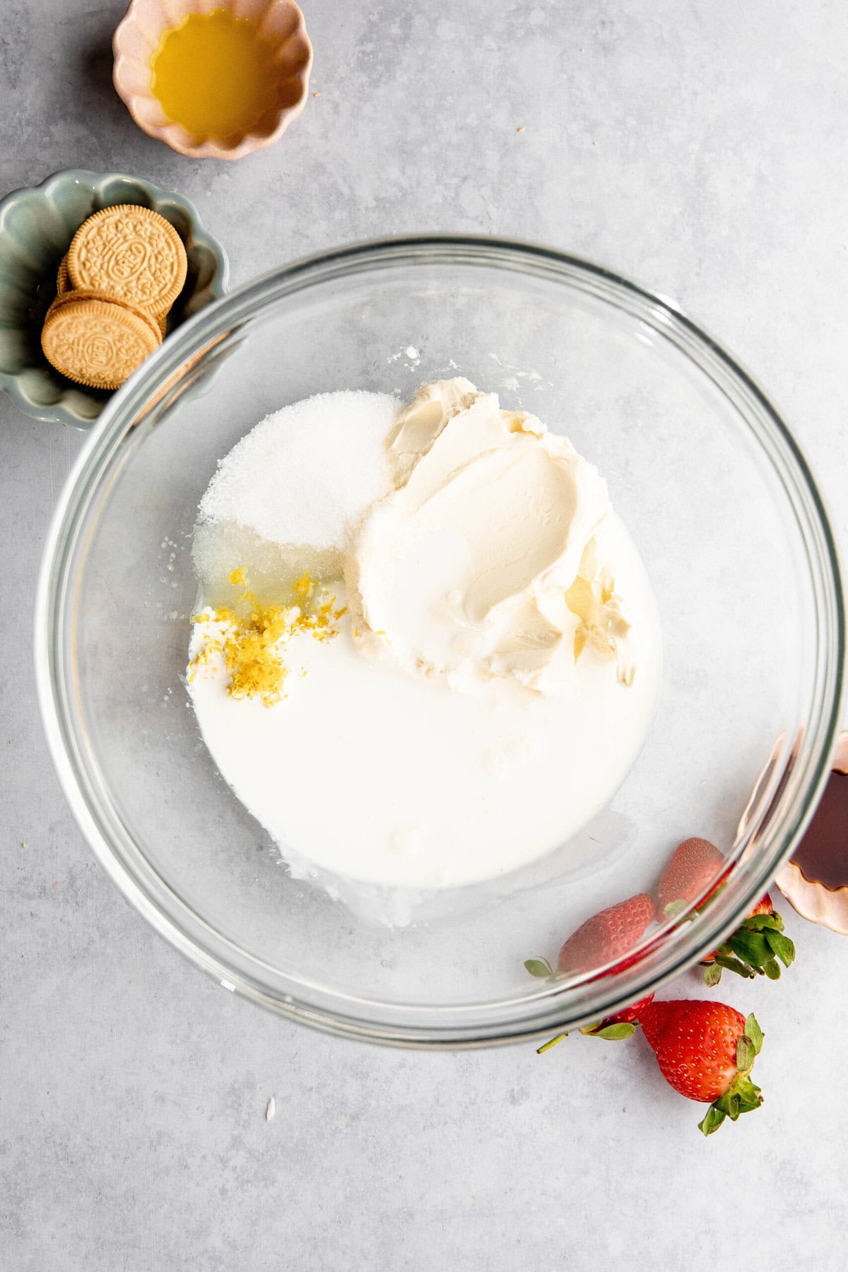 A clear glass bowl containing cream cheese, sugar, lemon zest, and cream evokes thoughts of a delightful lemon lasagna. Strawberries, vanilla extract, golden sandwich cookies, and a small bowl of melted butter are nearby on a gray surface.