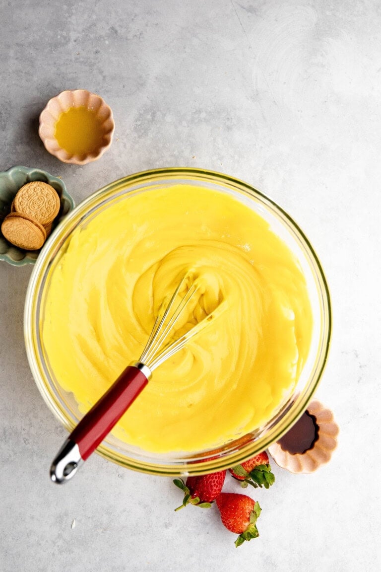 A glass bowl filled with yellow custard, reminiscent of a delicious lemon lush, has a whisk inside. Surrounding the bowl are small dishes with orange liquid, cookies, strawberries, and vanilla extract.