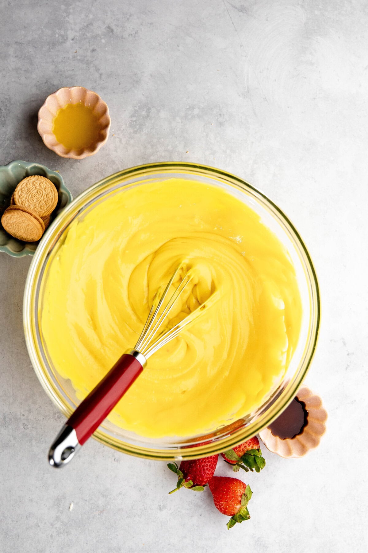 A glass bowl filled with yellow custard, reminiscent of a delicious lemon lush, has a whisk inside. Surrounding the bowl are small dishes with orange liquid, cookies, strawberries, and vanilla extract.