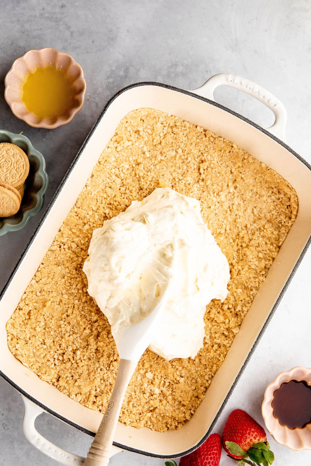 A rectangular baking dish with a crumbly base topped with a large dollop of cream mixture, resembling a delightful lemon lush. Nearby are small bowls with cookies, melted butter, and strawberries. A wooden spoon is in the dish.