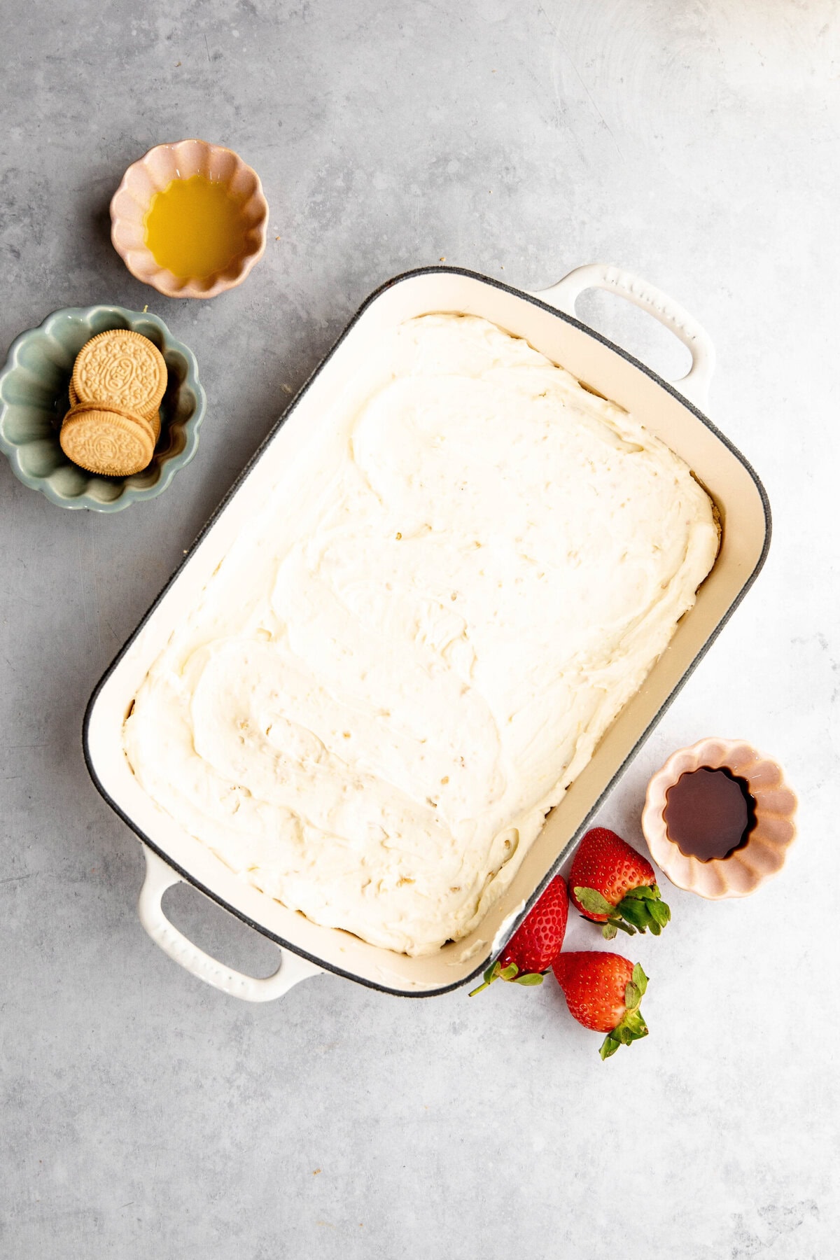 A rectangular baking dish filled with a creamy lemon lush mixture is placed on a surface. Surrounding it are small bowls with oil, cookies, strawberries, and a liquid, possibly vanilla extract.