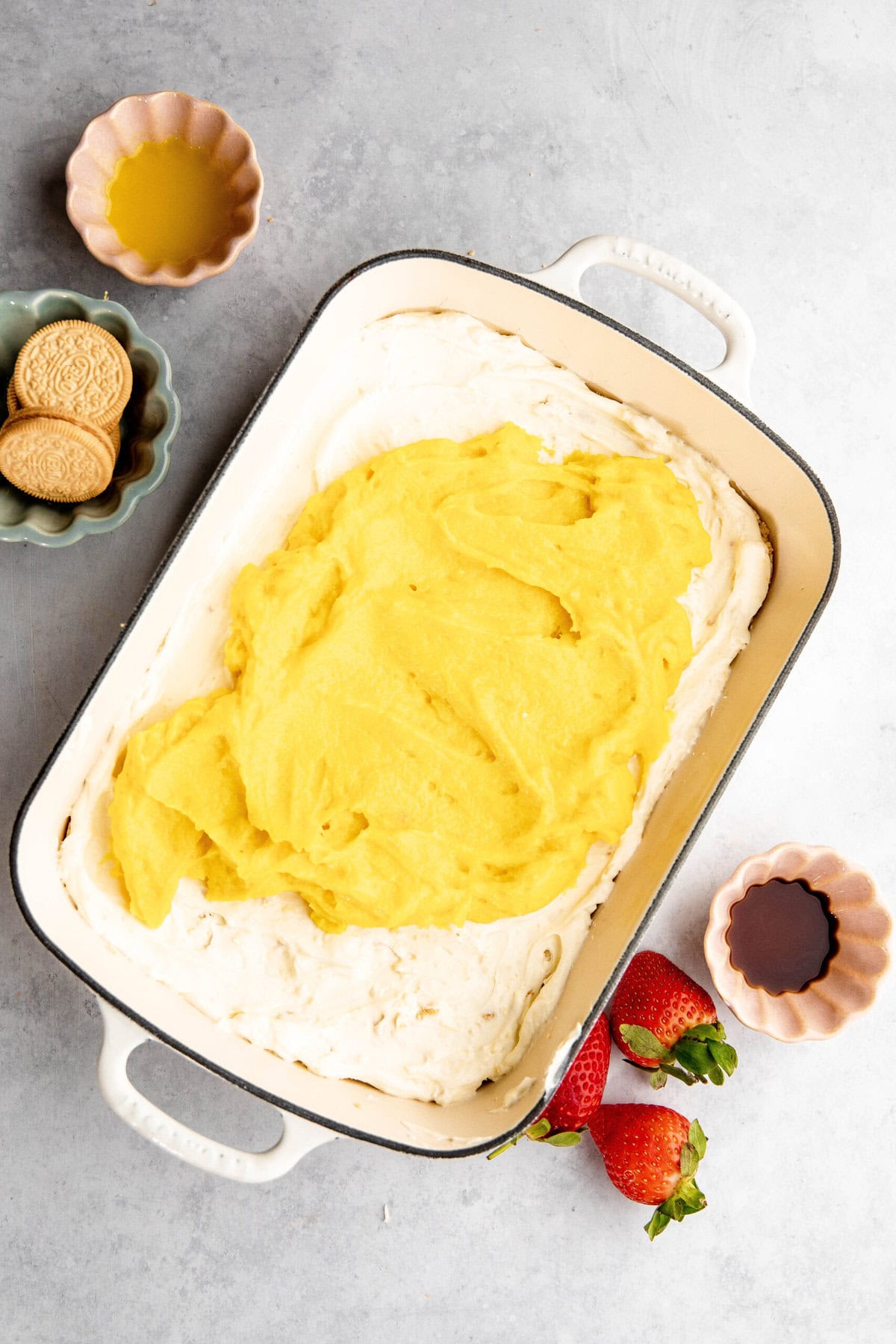 A baking dish with a layer of cream and a yellow mixture, reminiscent of lemon lasagna. Surrounding it are strawberries, two small bowls with liquids, and another bowl with cookies.