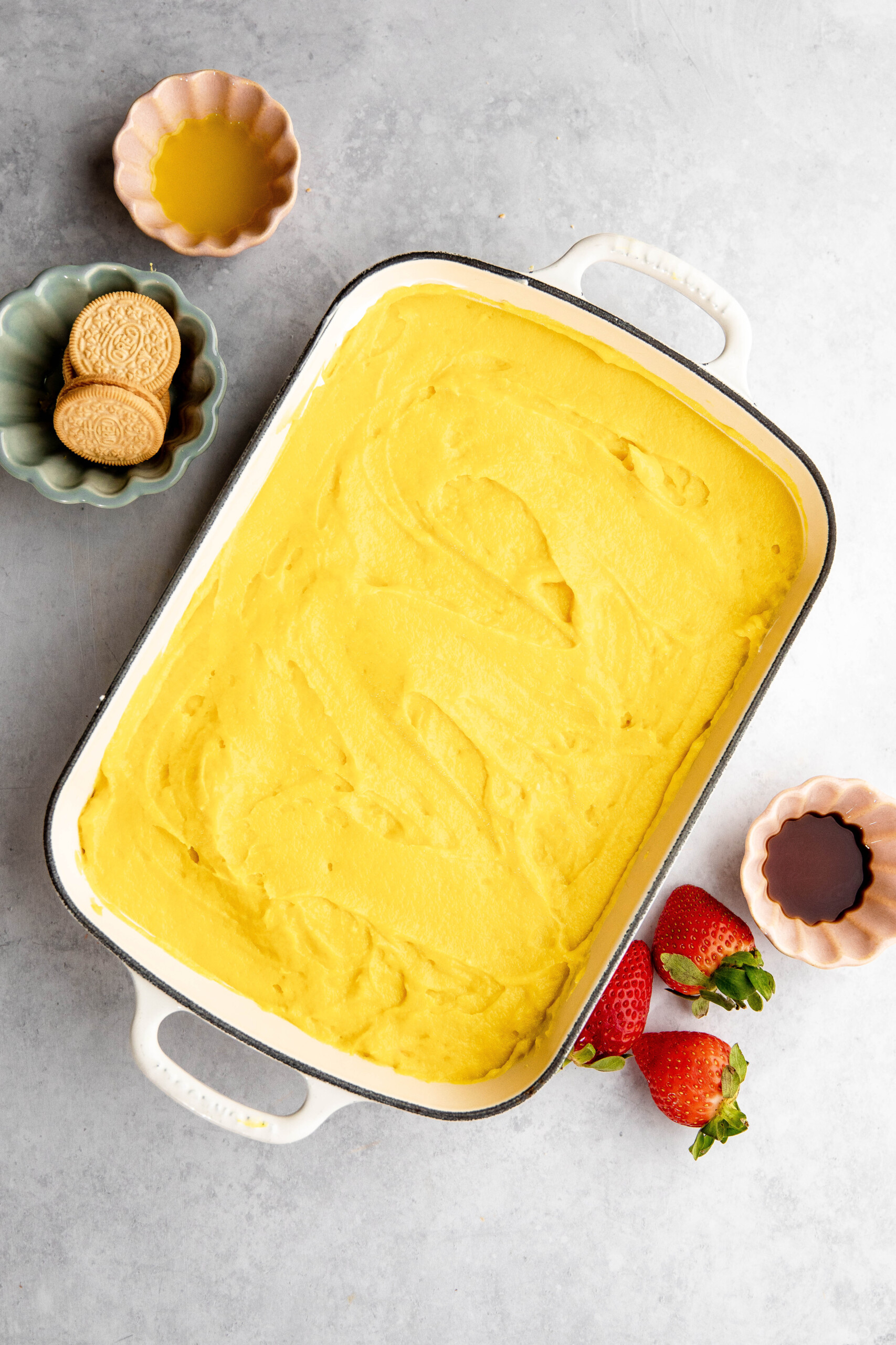 A baking dish filled with yellow batter sits next to small bowls containing vanilla wafers, liquid ingredients, and strawberries, hinting at a delightful lemon lush recipe in progress.