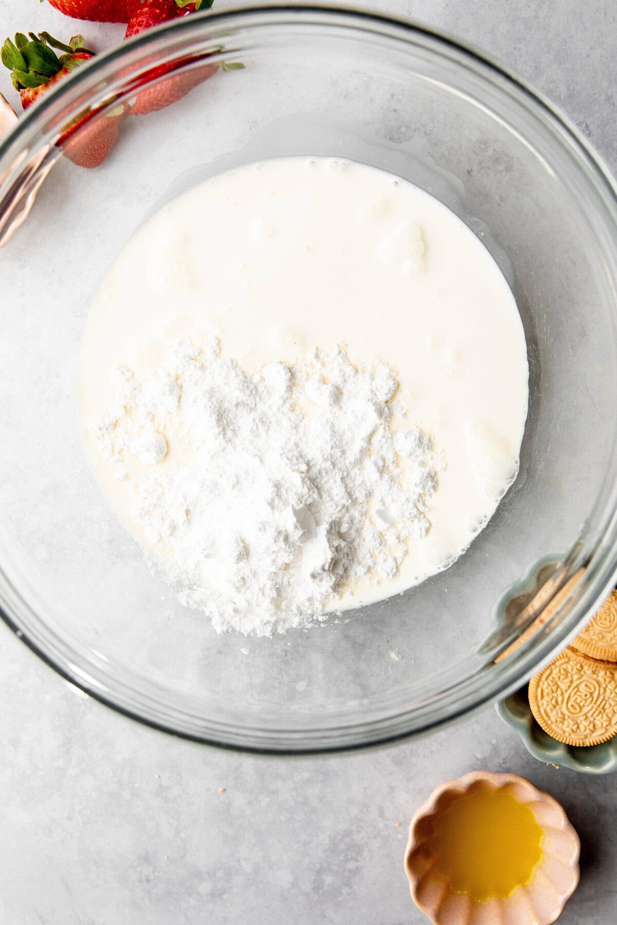 A glass bowl filled with a mixture of cream and powdered ingredients sits on a table, surrounded by strawberries, cookies, and melted butter in separate bowls—perfect for creating a delightful lemon lush dessert.