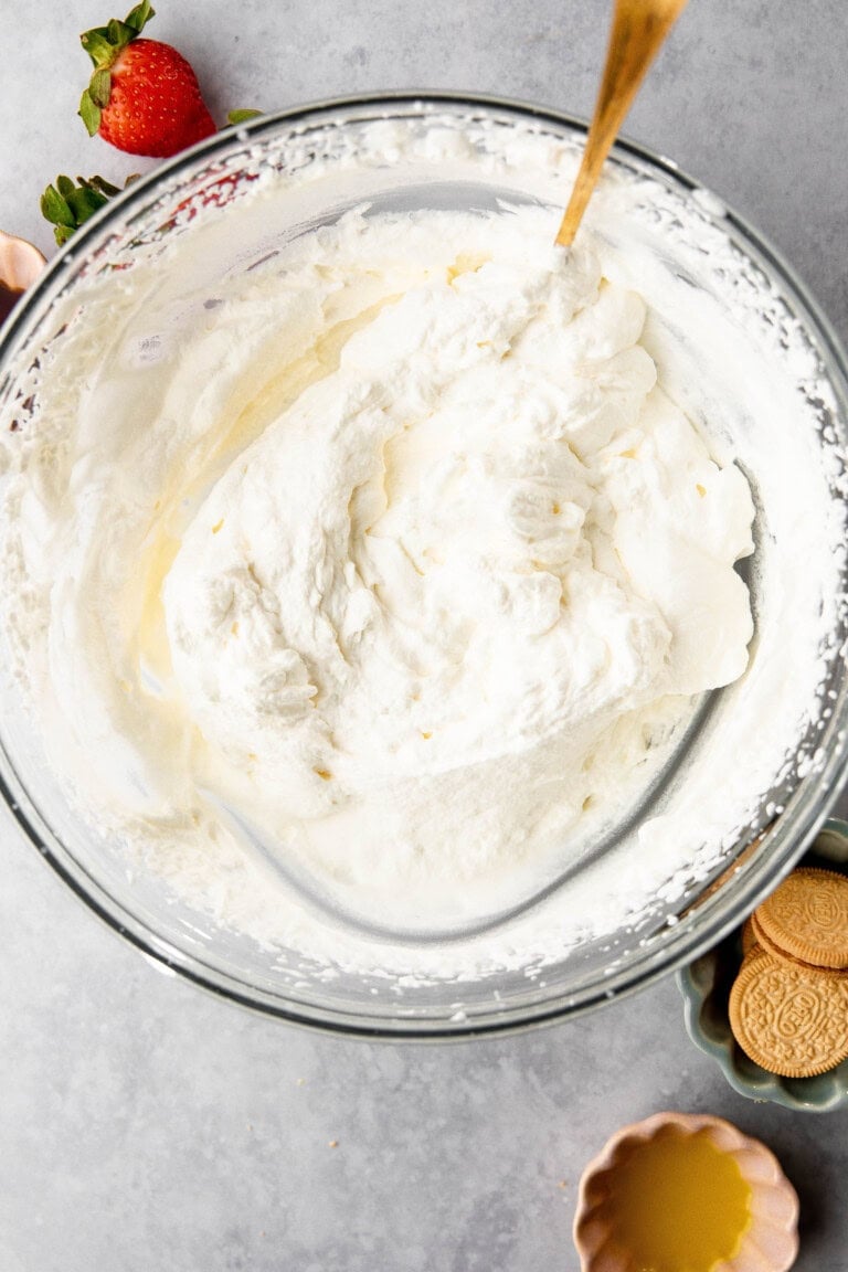 A clear glass bowl filled with whipped cream sits on a gray surface. Nearby are a strawberry, cookies, and a small bowl of yellow liquid reminiscent of lemon lush. A wooden spoon is in the whipped cream.