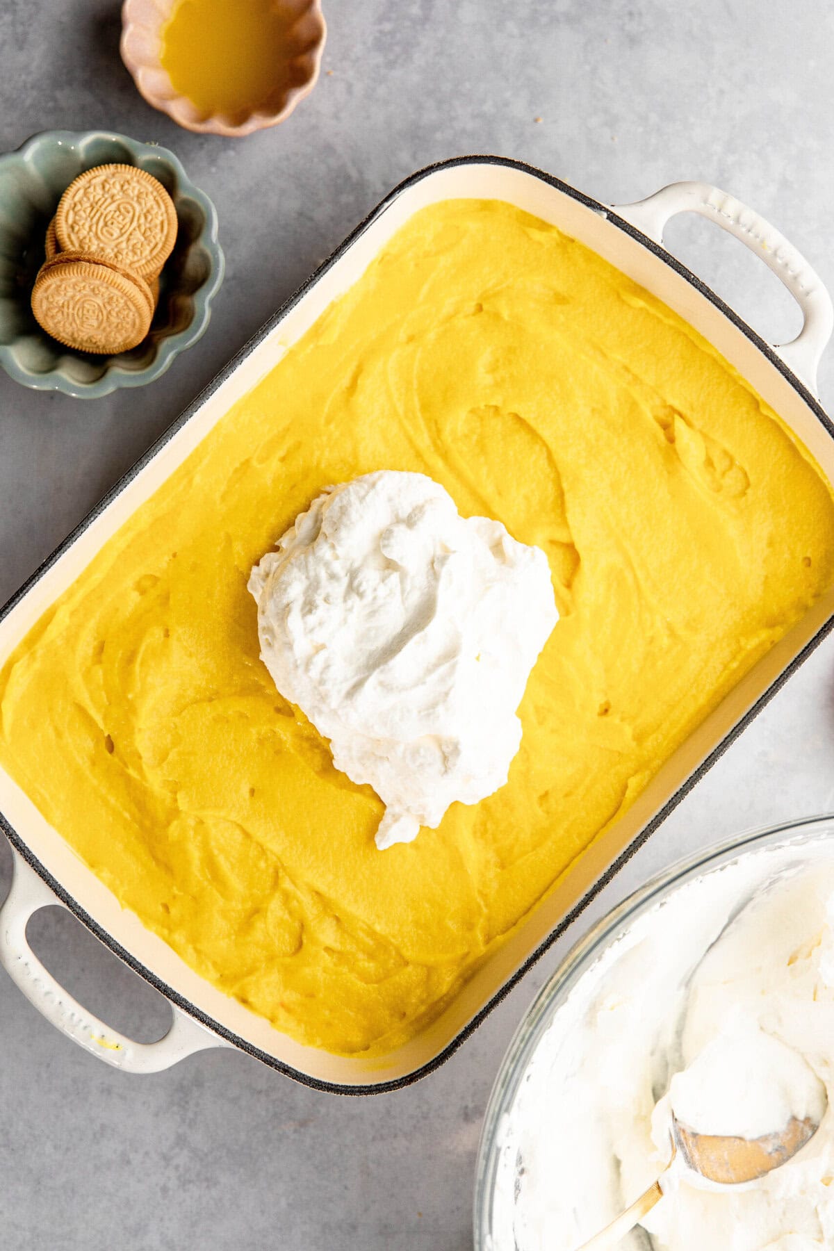 A baking dish filled with a layer of bright yellow pudding, reminiscent of lemon lush, topped with a dollop of whipped cream. Surrounding the dish are a bowl of cream, a small dish of cookies, and a cup of honey.