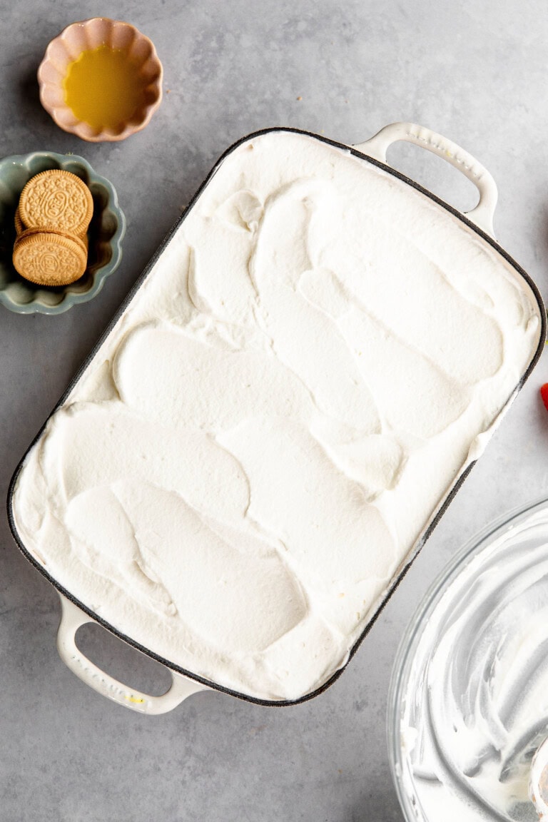 A baking dish filled with a layer of whipped cream sits on the countertop, evoking memories of a delicious lemon lush. Nearby, there is a bowl of golden cookies and a small dish of yellow liquid.