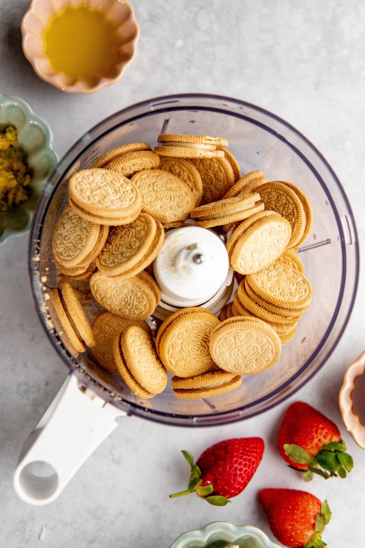 A food processor filled with vanilla sandwich cookies, surrounded by small bowls containing melted butter and strawberries on a gray countertop, ready to be transformed into a delightful lemon lasagna.