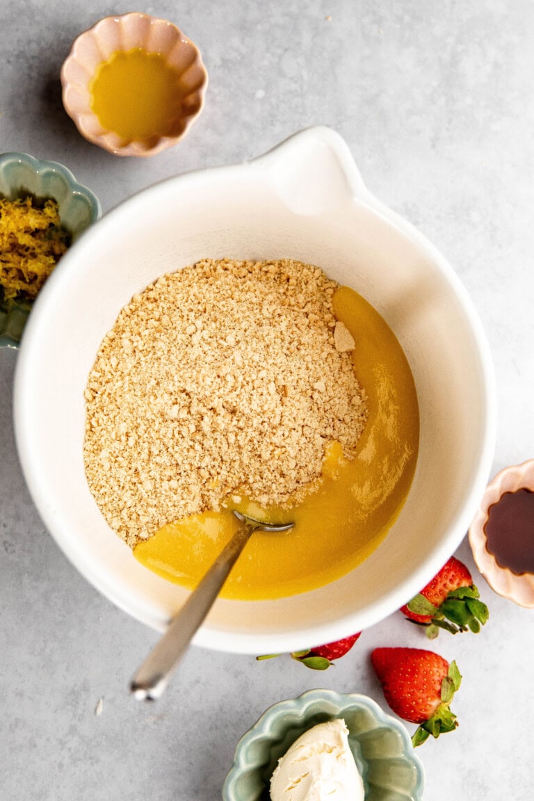 Top-down view of a mixing bowl containing a crumb mixture and melted butter, perfect for creating a base layer for lemon lasagna. Surrounding the bowl are small dishes with various ingredients, including lemon zest, strawberries, and an unknown liquid.