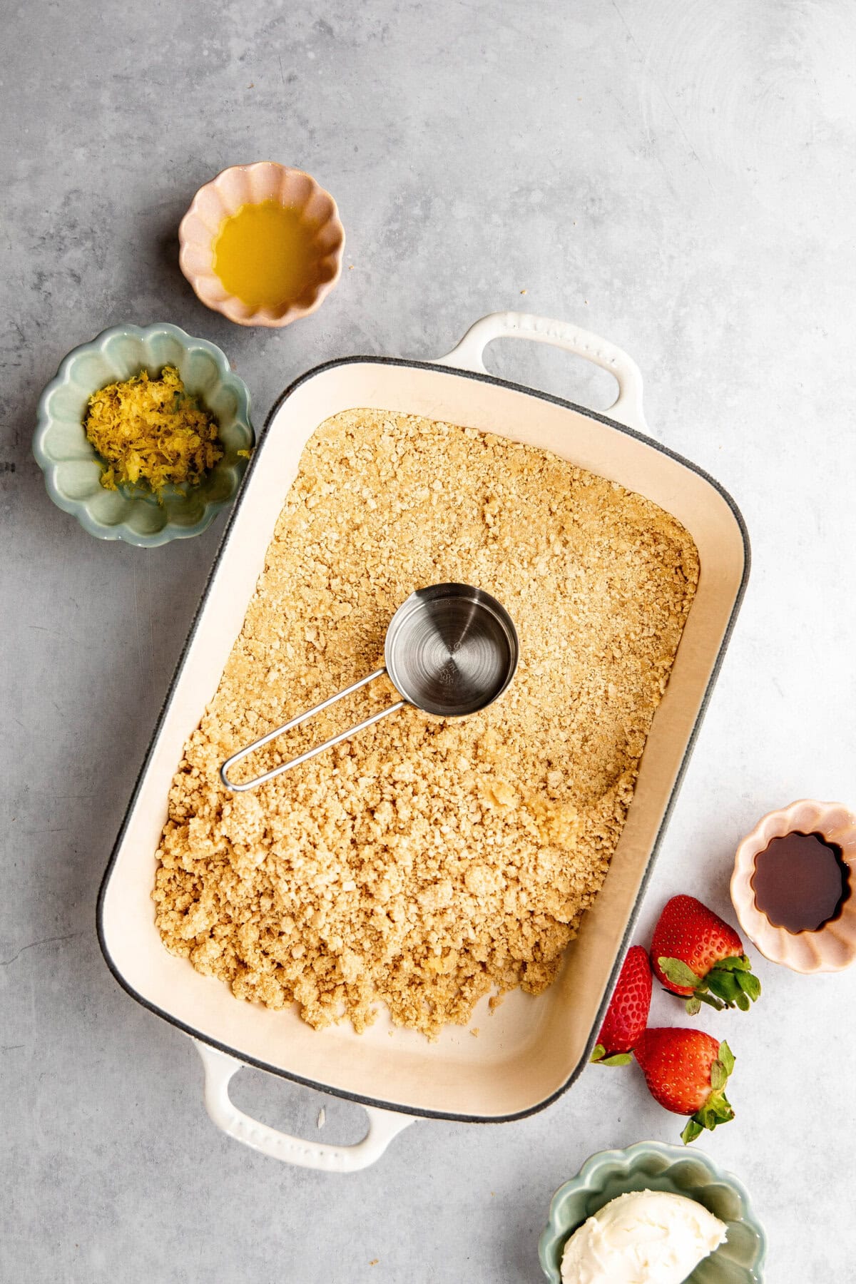 A baking dish with a crumbly mixture and a measuring cup sits surrounded by small bowls of ingredients: lemon zest, melted butter, vanilla extract, strawberries, and white cream. This is the beginning of a delectable lemon lush dessert, where each layer promises tangy and sweet perfection.