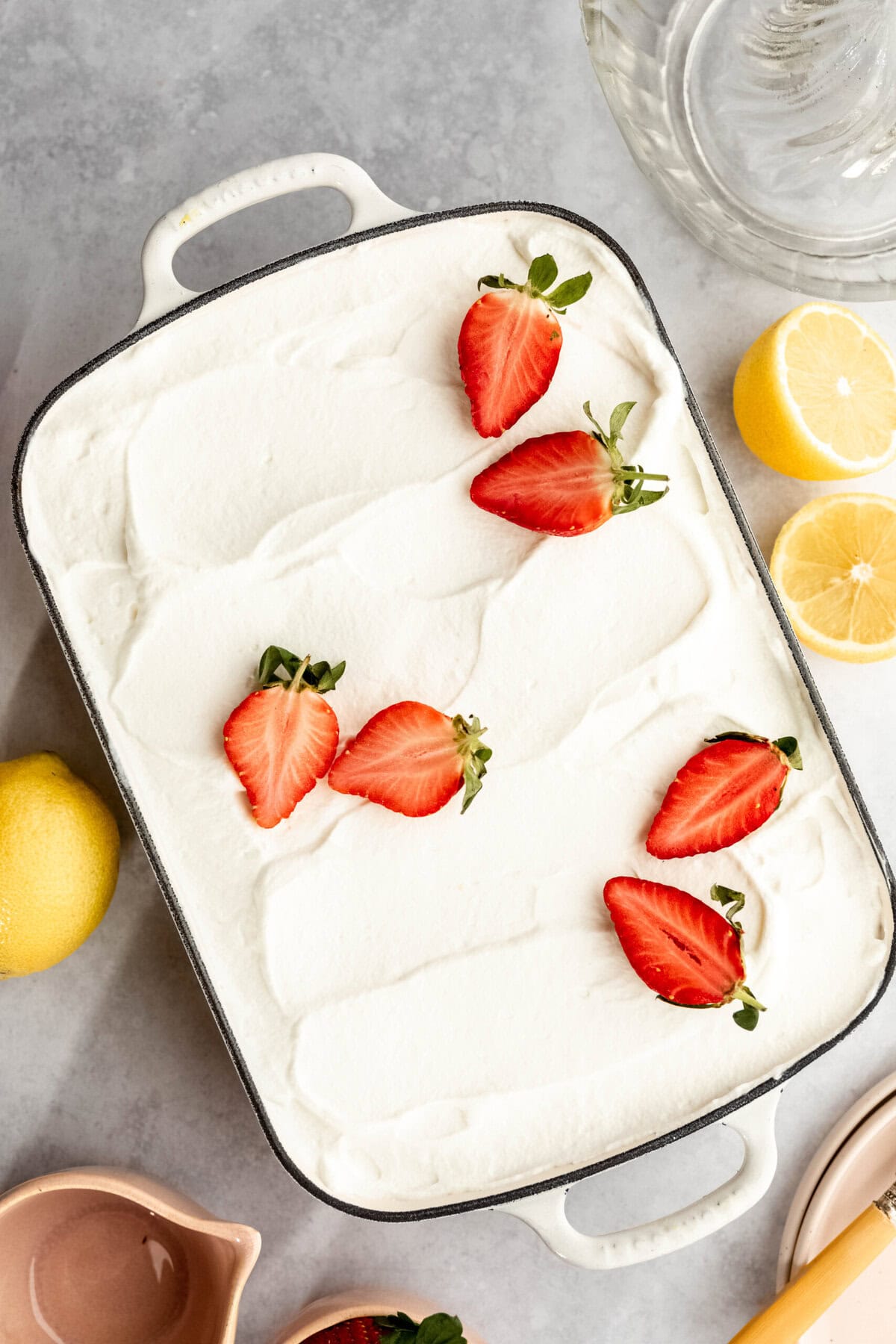 A dessert dish with a layer of white cream, topped with five sliced strawberries, is surrounded by lemons and various preparation tools on a grey countertop.