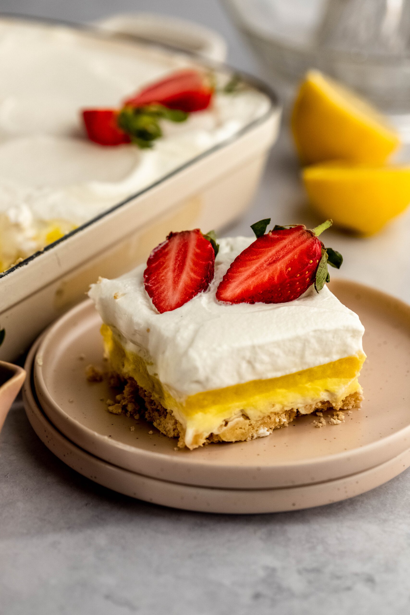 A slice of layered dessert with a golden oreo crust, yellow filling, and whipped cream topping, garnished with sliced strawberries, on a beige plate. The remaining dessert is in a baking dish nearby.