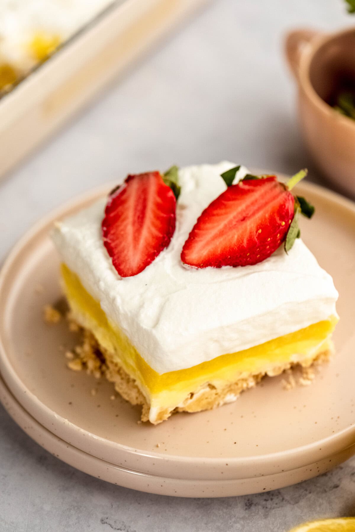 A slice of layered dessert with a crumbly base, yellow custard filling, whipped cream topping, and two strawberry slices on a beige plate.