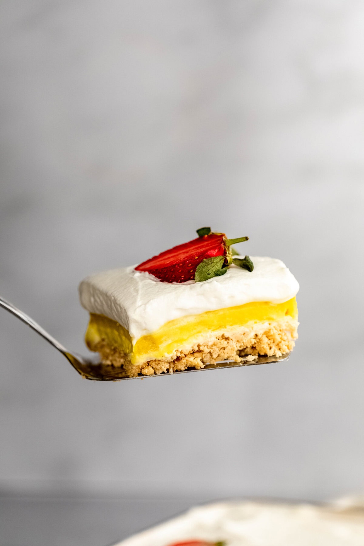 A slice of layered dessert with a crumbly base, yellow filling, white whipped topping, and a sliced strawberry on top, held on a fork against a neutral background.