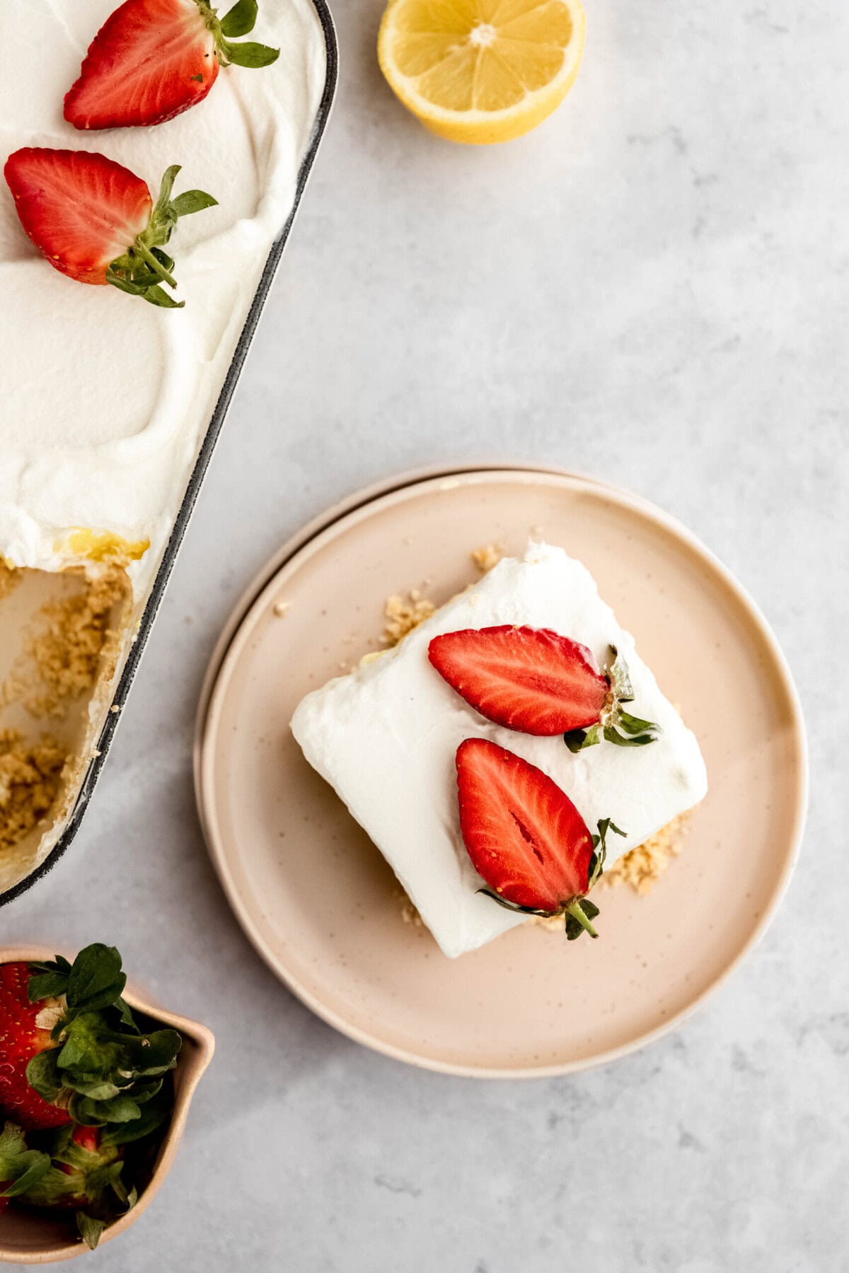 A rectangular slice of creamy dessert topped with two strawberry halves on a small, round plate beside a baking dish with more servings. A lemon half and a container of strawberries are nearby.