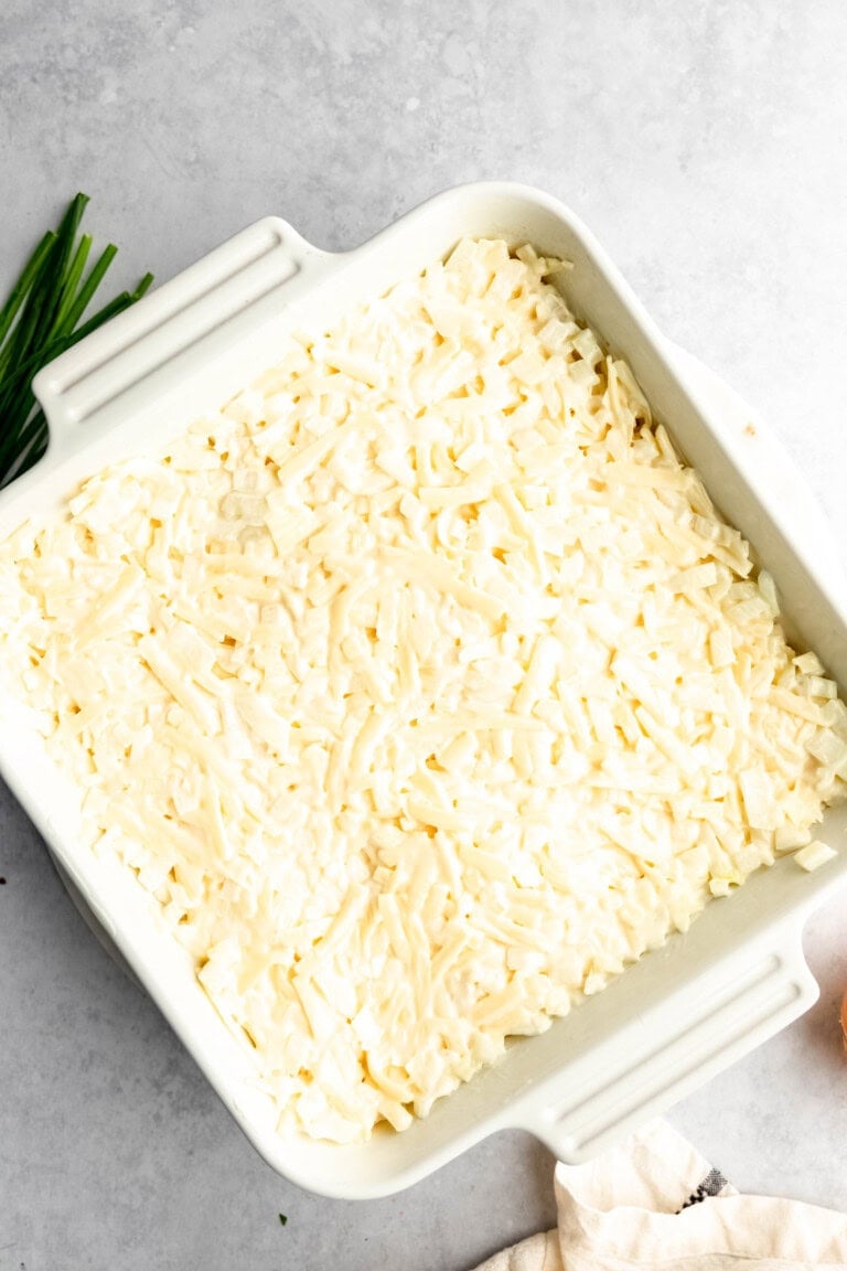 A white rectangular baking dish filled with grated cheese or a similar food topping, placed on a light gray surface with some green herbs partially visible in the background.