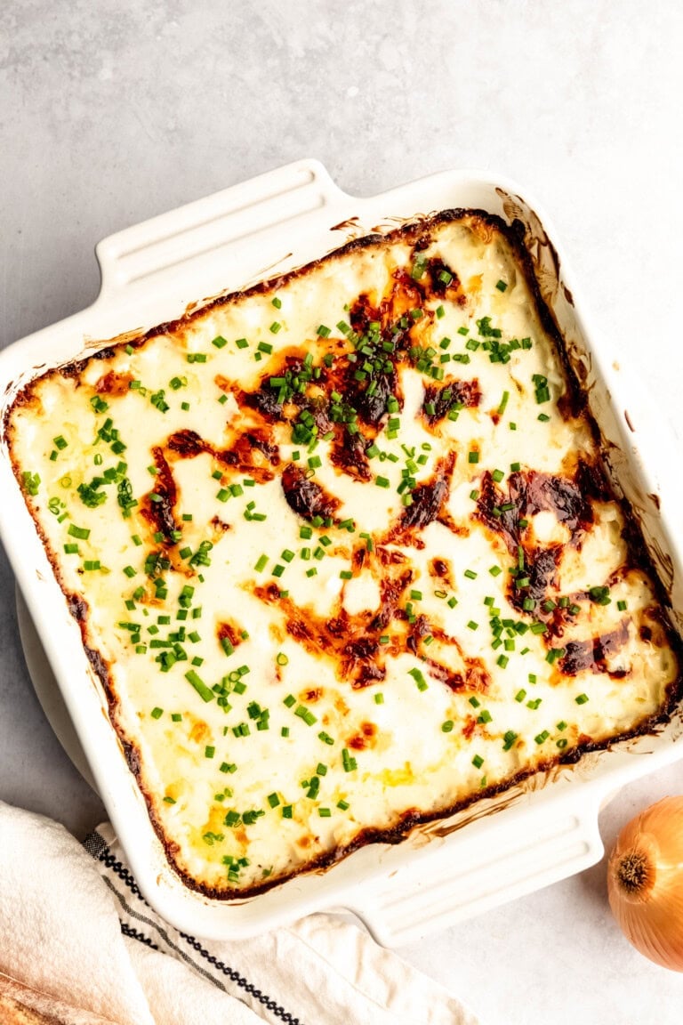 A baked, creamy casserole topped with melted cheese and sprinkled with chopped green herbs in a white square dish. An onion and a kitchen towel are placed to the side.