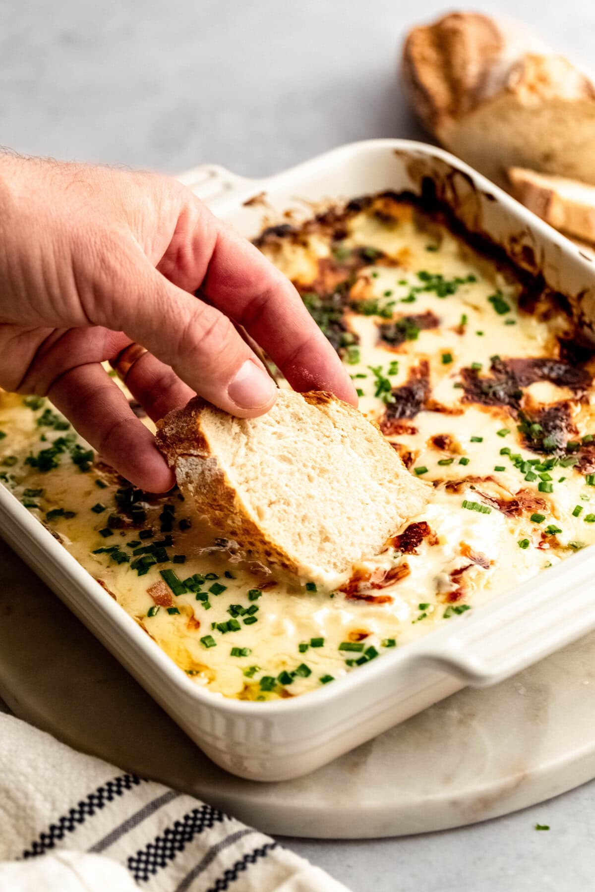 A hand dipping a piece of bread into a casserole dish filled with a creamy baked dip topped with chives and browned bits. There is a cloth napkin and a loaf of bread in the background.